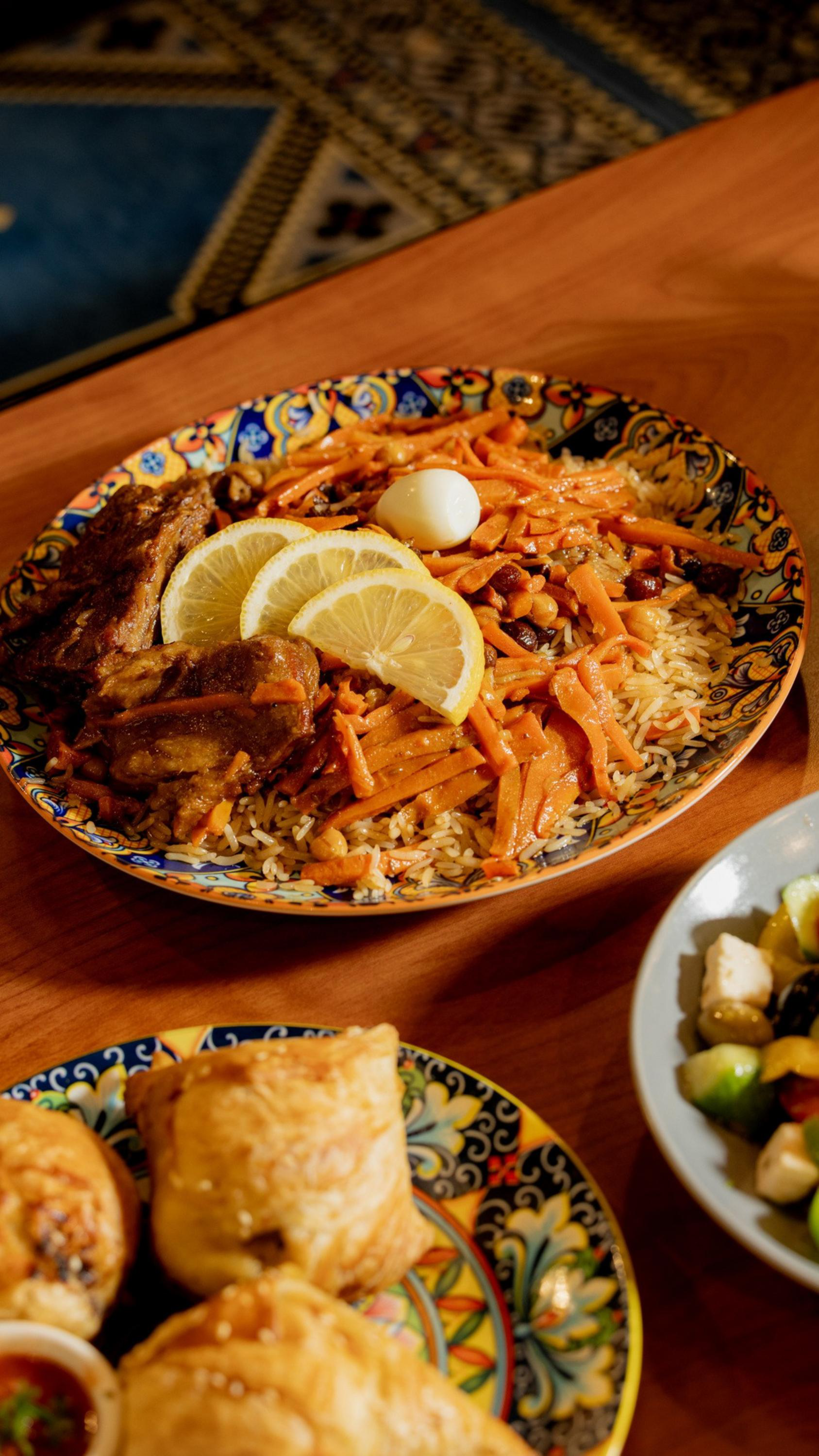 A colorful plate holds rice topped with cooked carrots, three lemon slices, a boiled egg, and pieces of meat. Another plate contains pastries.