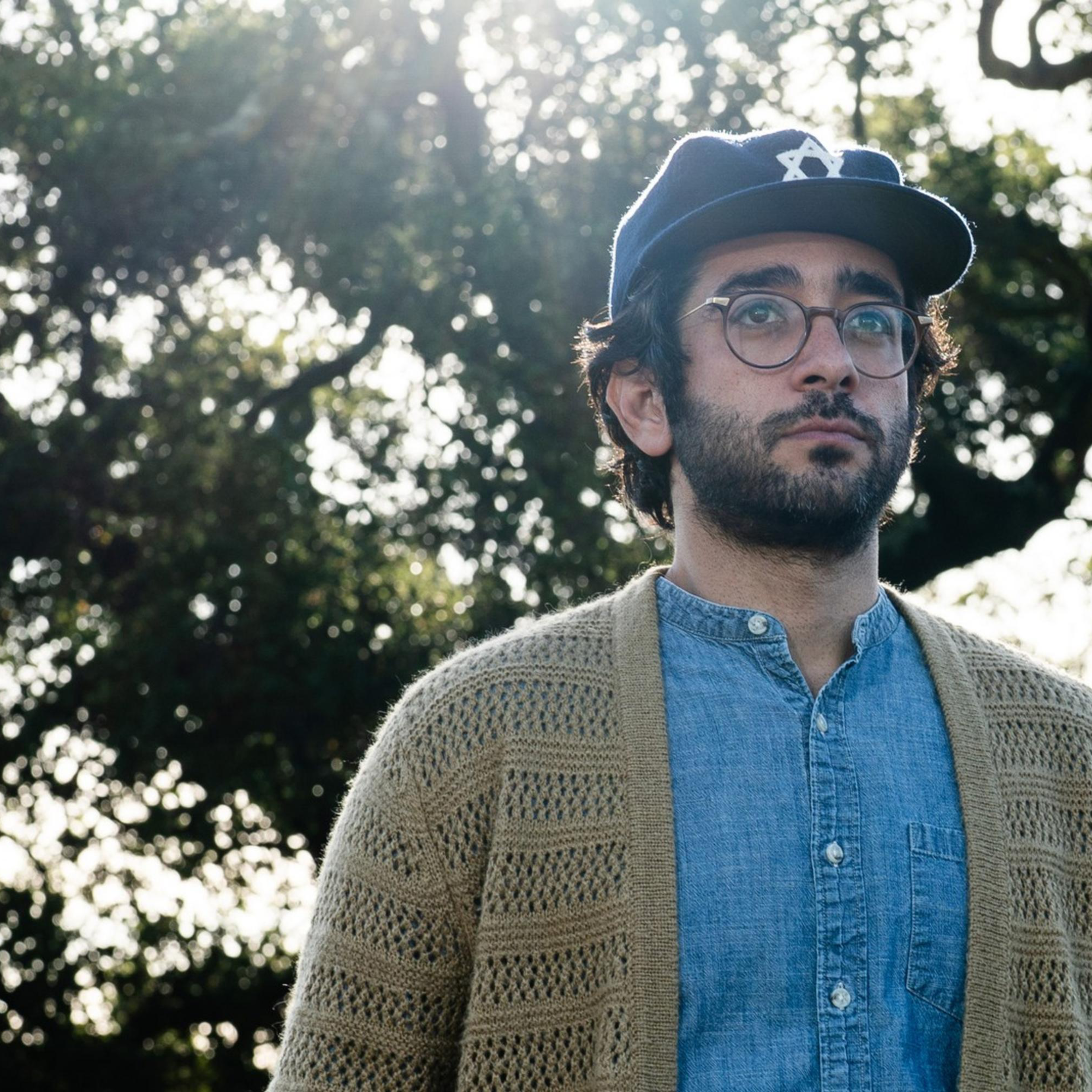 A person with a beard, glasses, and baseball cap stands outdoors. They wear a blue shirt and knitted cardigan. Sunlight filters through trees in the background.