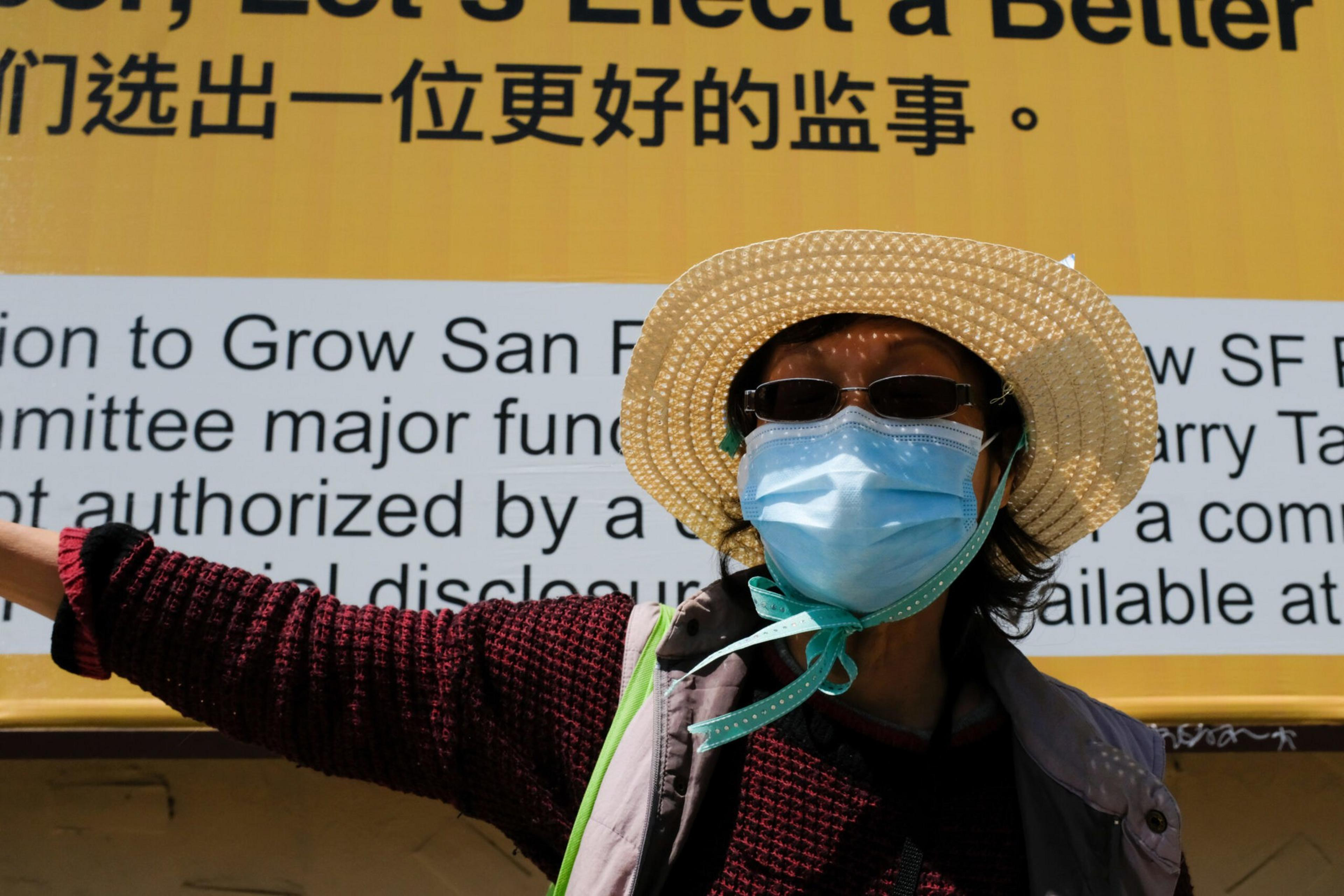 A person wearing a straw hat, sunglasses, and a face mask stands in front of a yellow sign with bilingual English and Chinese text.