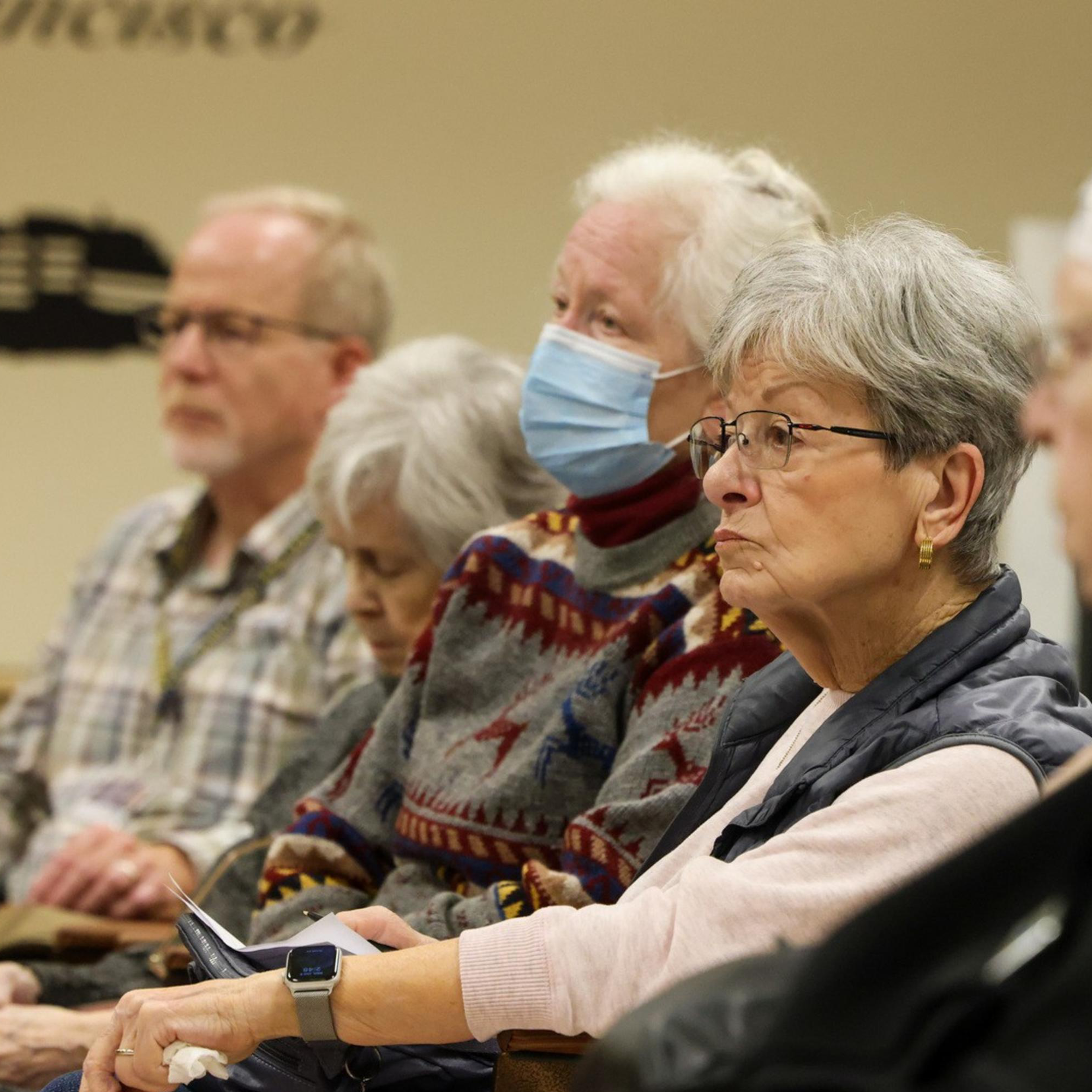 A group of elderly people, some wearing masks, sit attentively in a room. One person in a nun's habit is seen on the right. They appear to be listening intently.