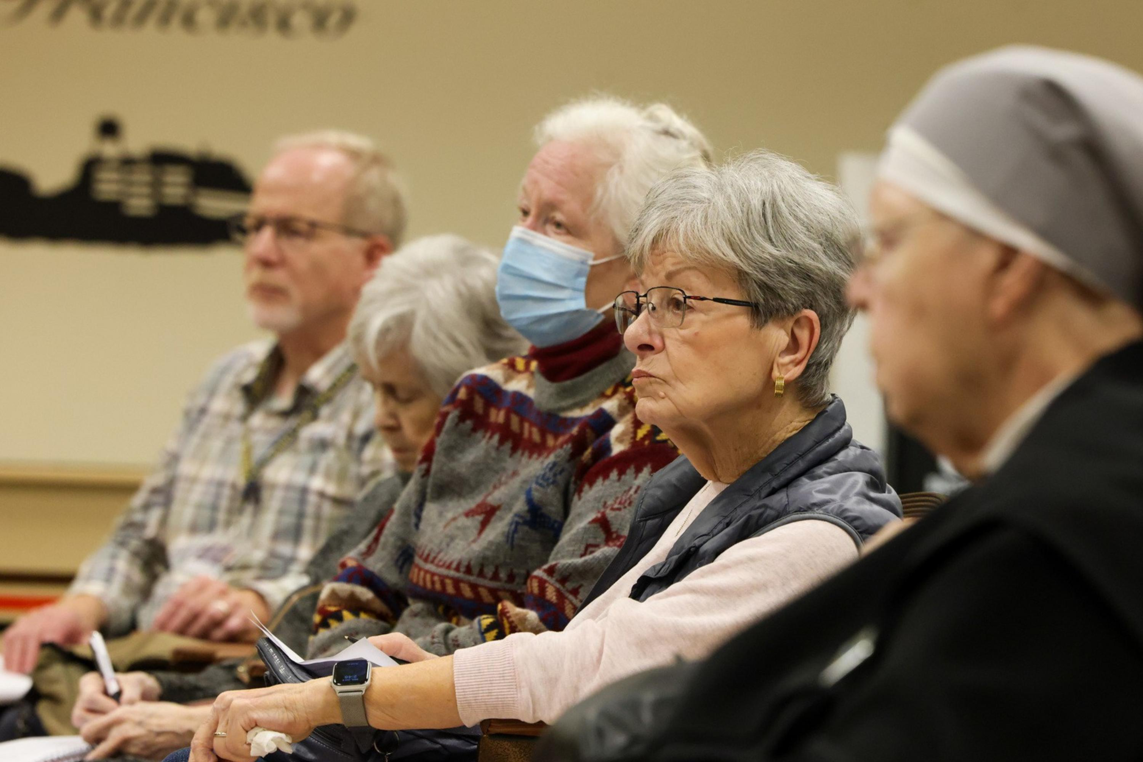 A group of elderly people, some wearing masks, sit attentively in a room. One person in a nun's habit is seen on the right. They appear to be listening intently.