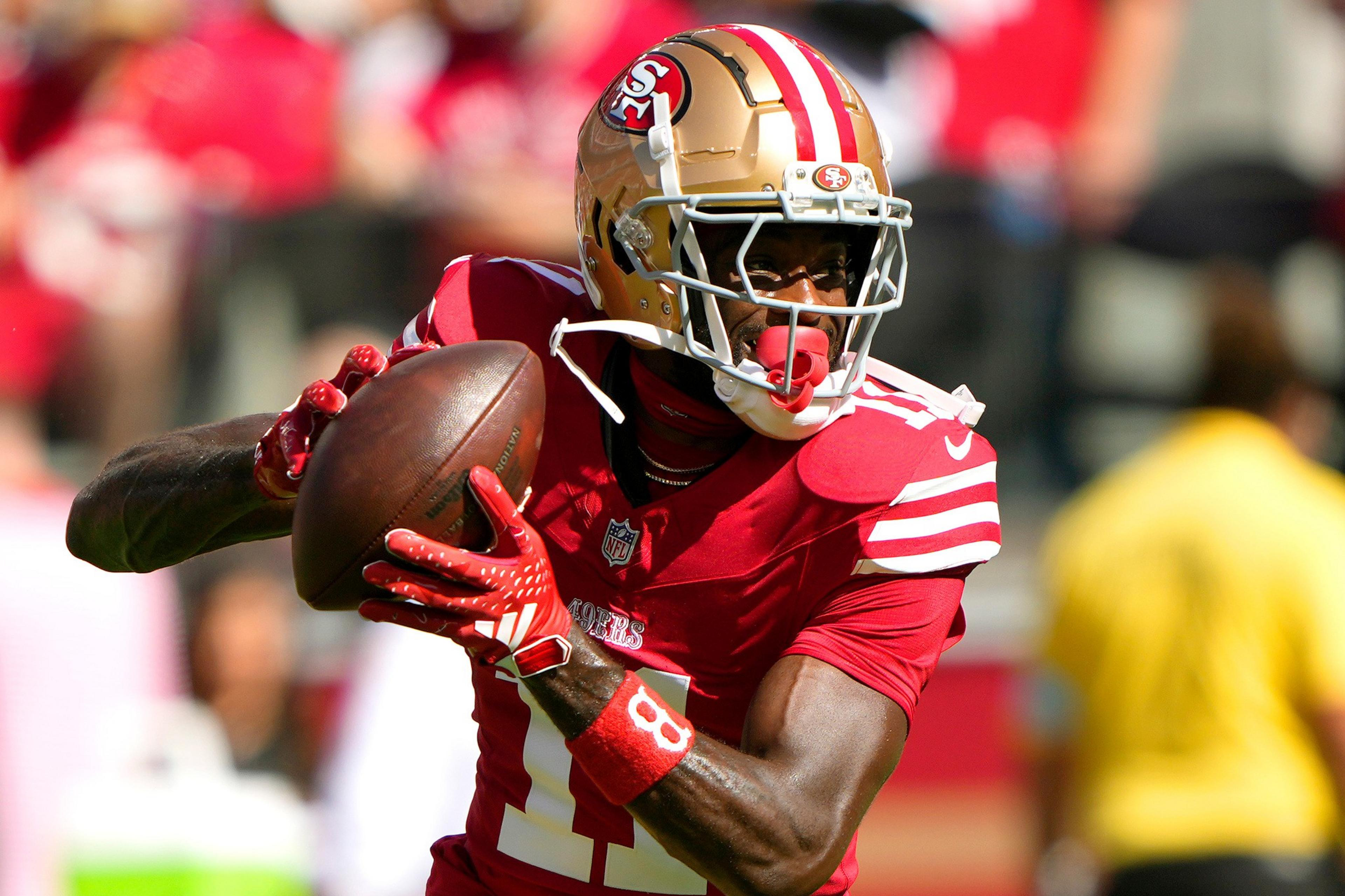 A San Francisco 49ers player in red and gold gear holds a football, looking to pass during a sunny game.