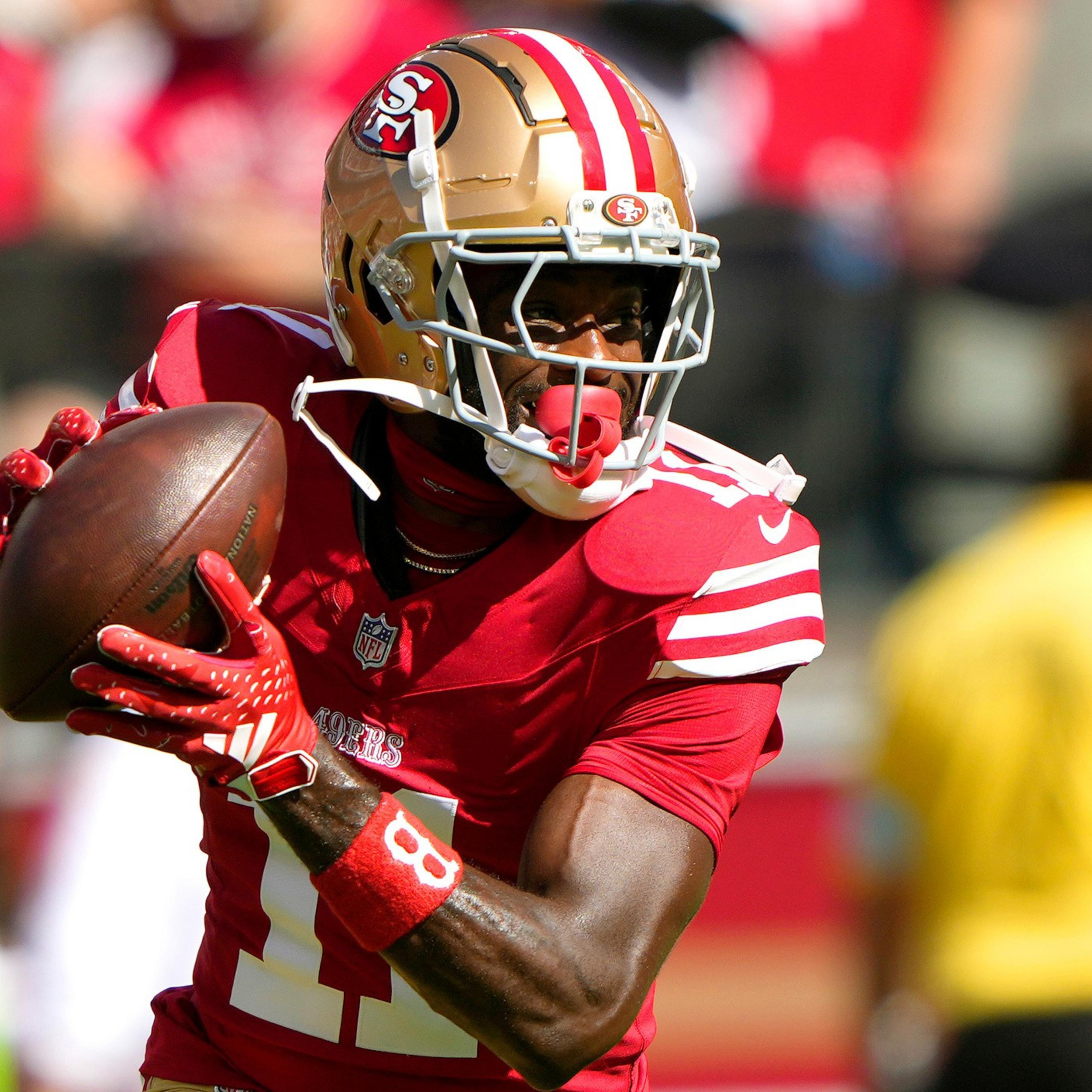 A San Francisco 49ers player in red and gold gear holds a football, looking to pass during a sunny game.