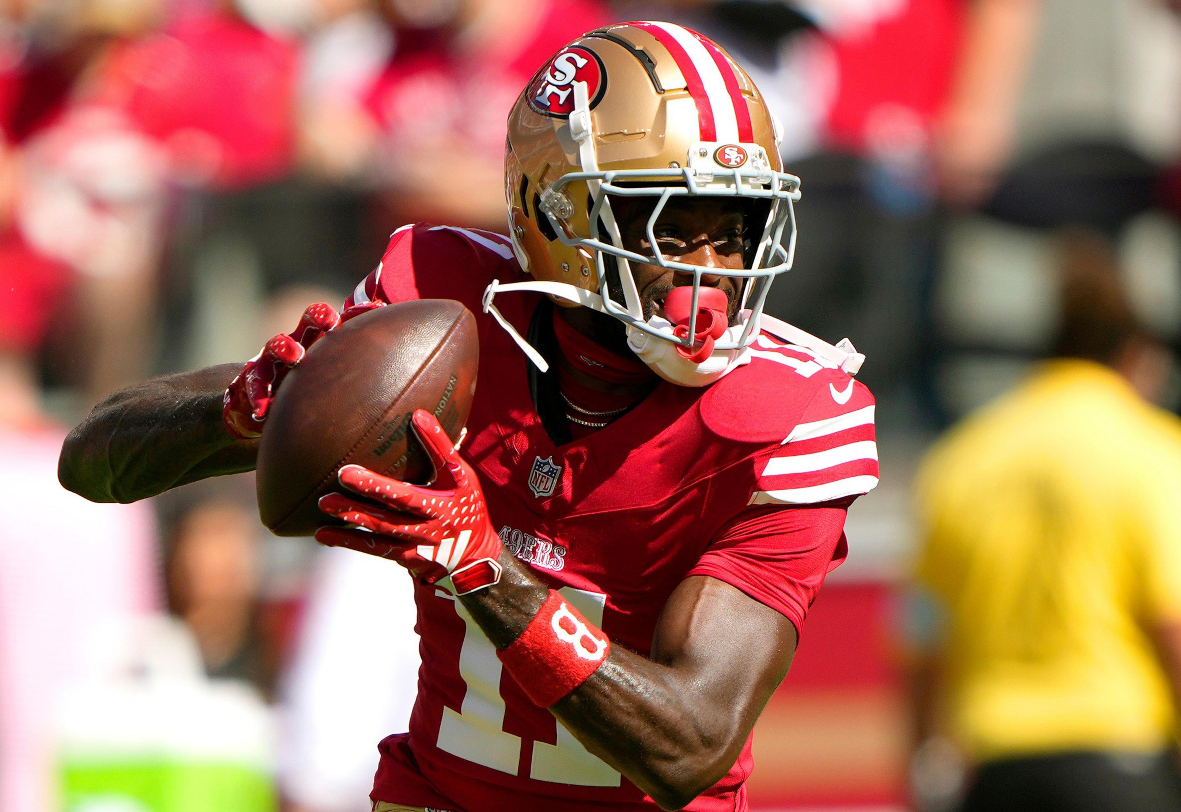 A San Francisco 49ers player in red and gold gear holds a football, looking to pass during a sunny game.