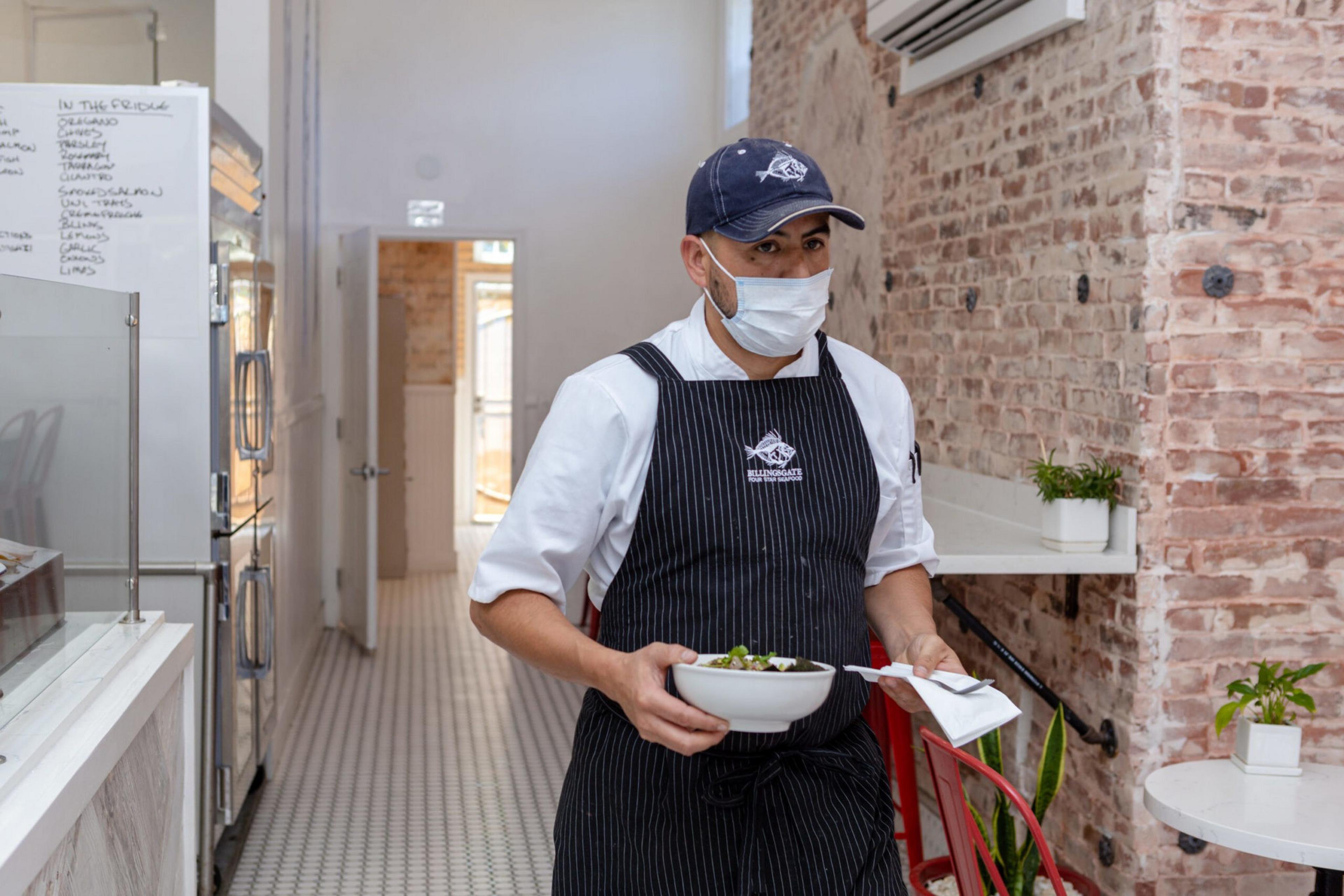 A chef wearing a mask, white shirt, black apron, and navy cap holds a bowl of food and a napkin while walking in a kitchen with exposed brick walls.