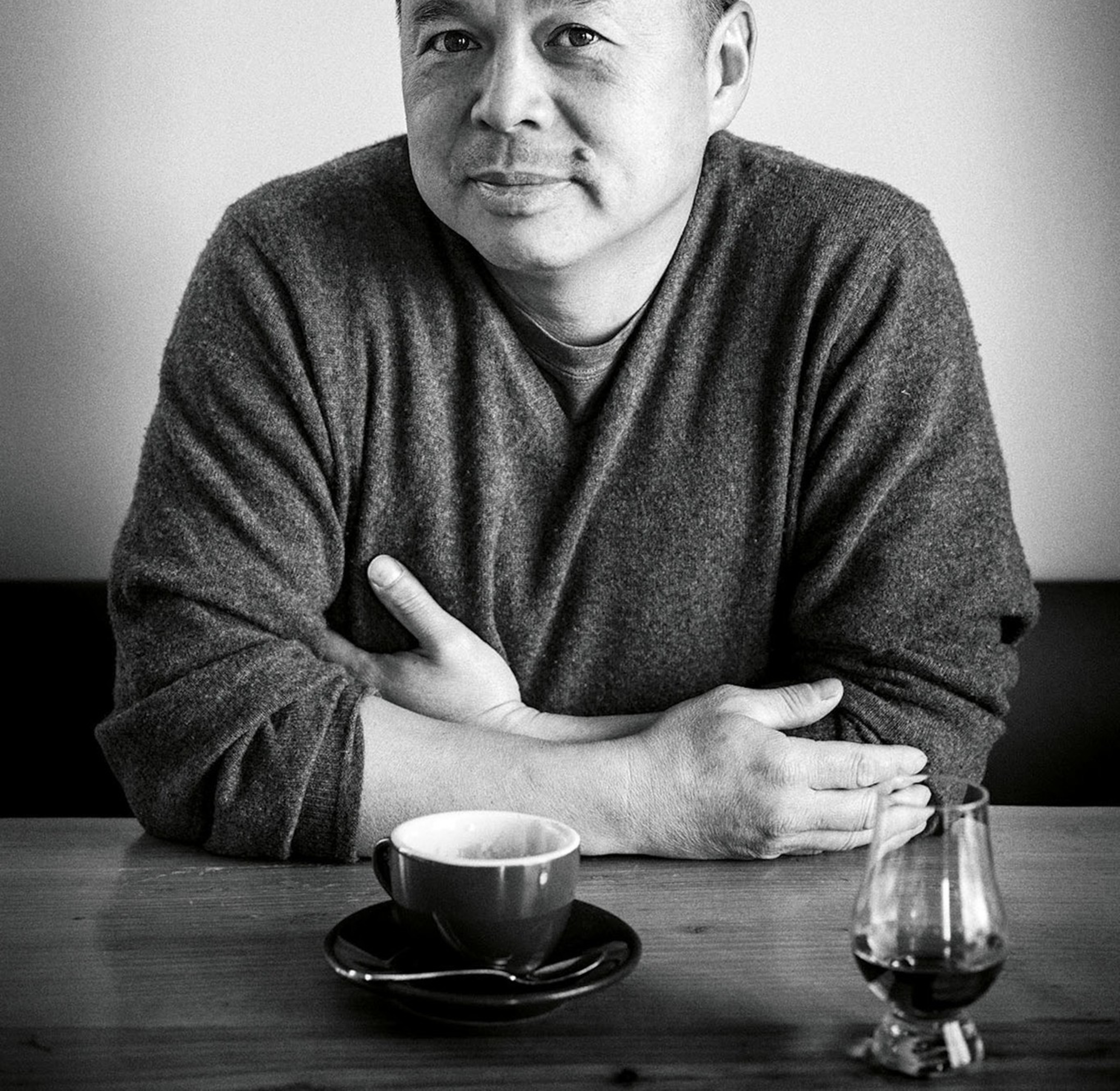 A man wearing a dark sweater sits at a table with a mug of coffee.