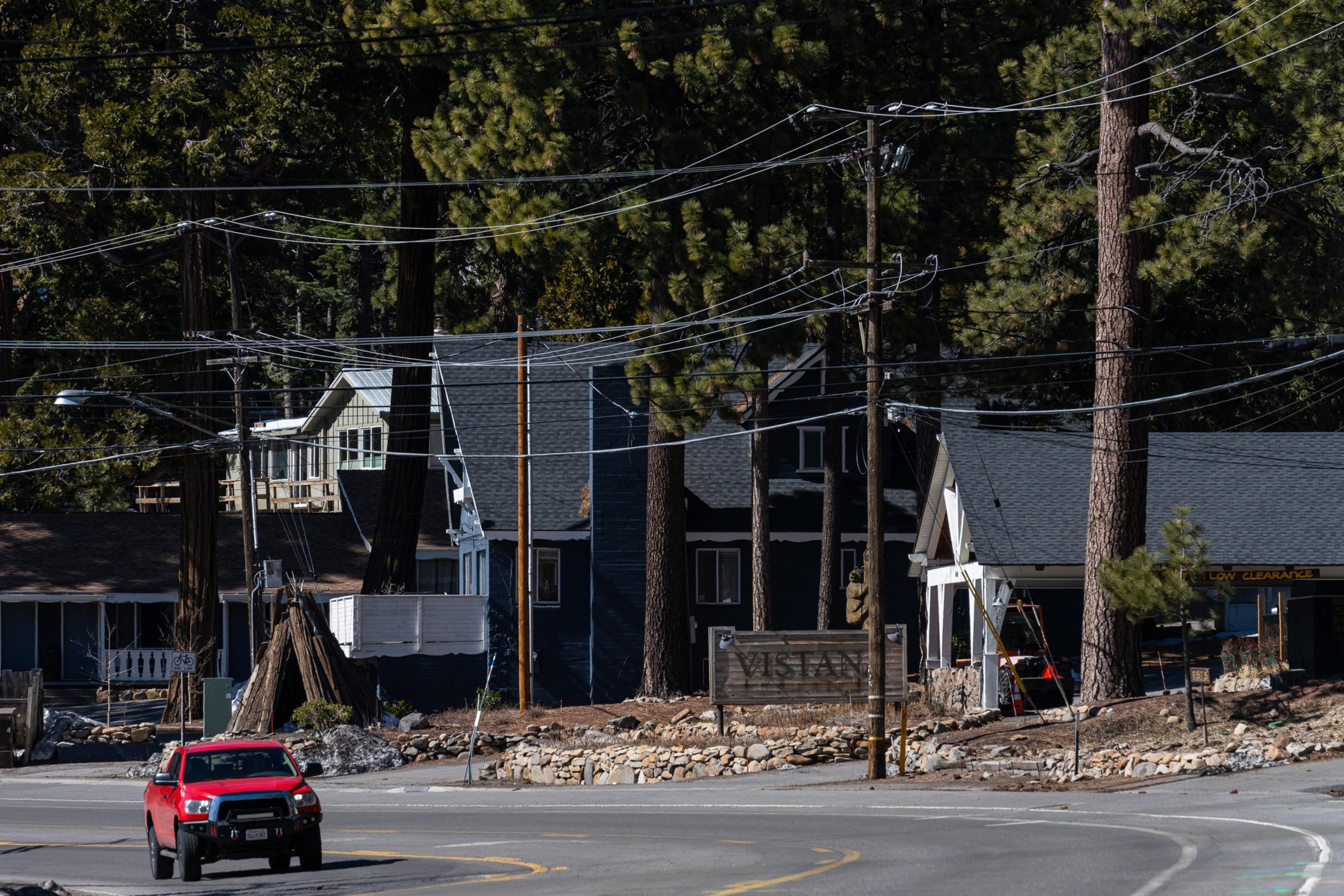 A red truck drives past a sign reading &quot;VISTAN&quot; in a wooded area with tall trees. There are houses surrounded by power lines and pine trees.