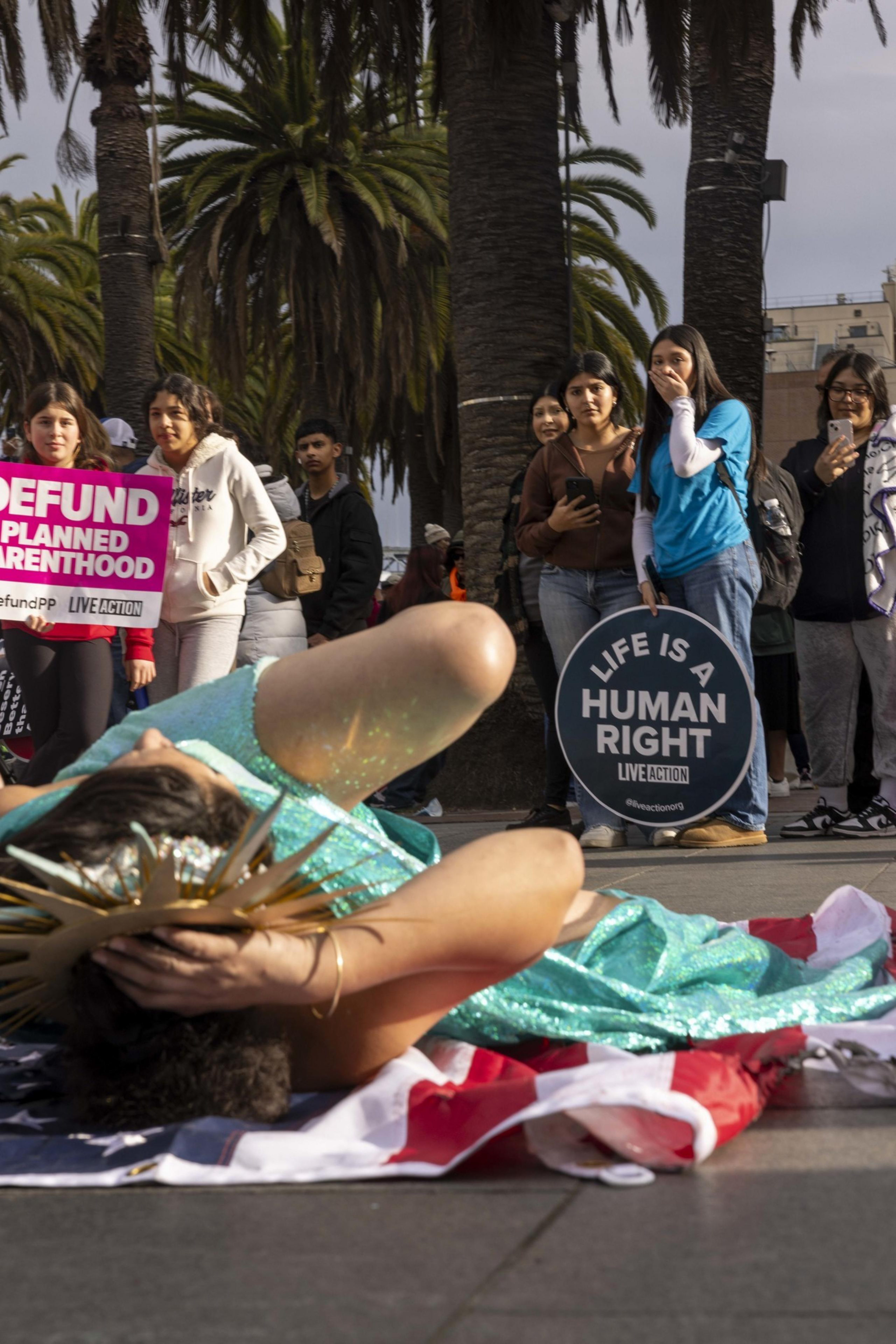 A person in a teal costume lies on an American flag, surrounded by people holding signs that read &quot;Defund Planned Parenthood&quot; and &quot;Life is a Human Right.&quot;