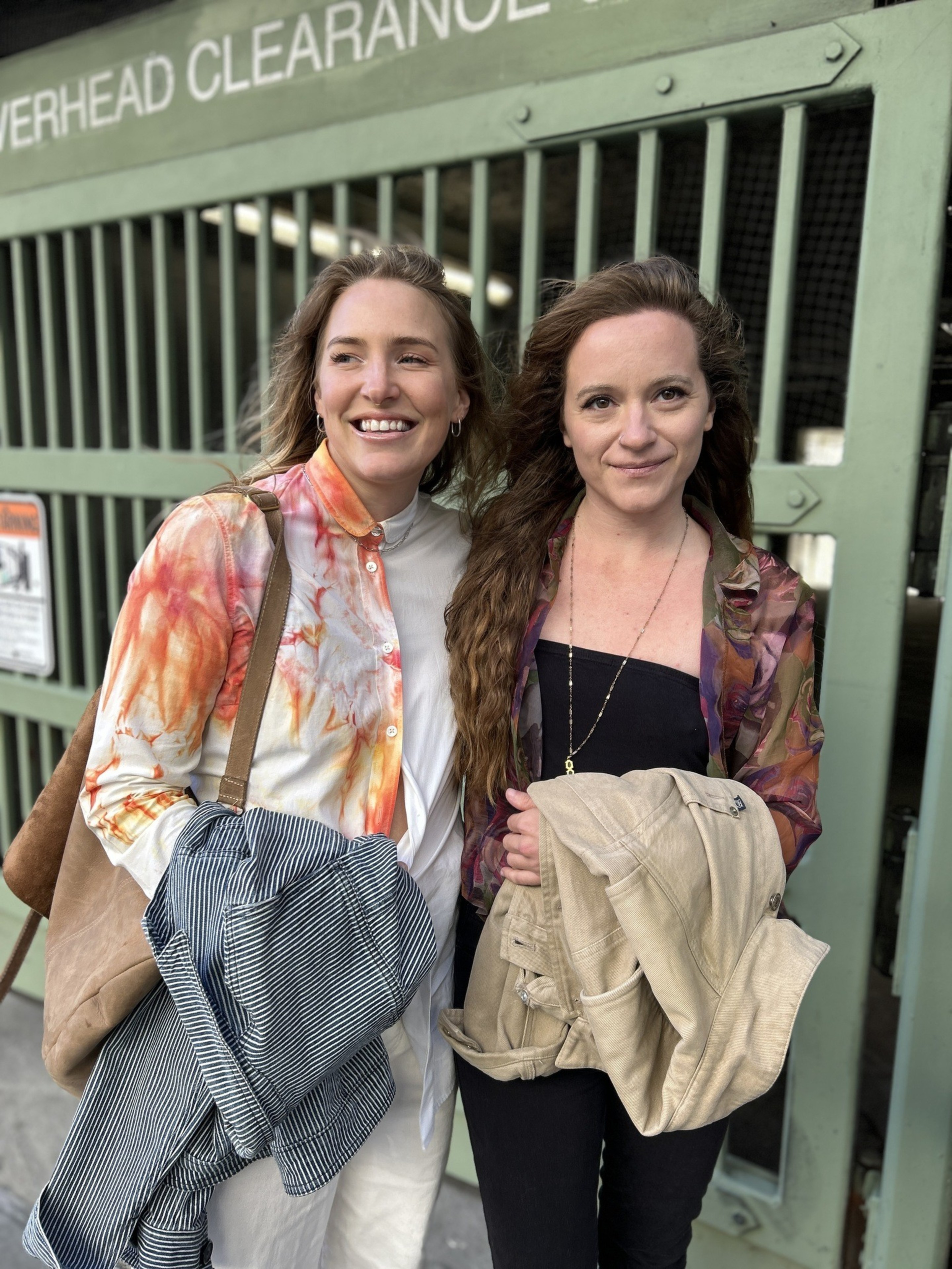 Two women smile and stand side by side, holding jackets. They are in front of a green barred gate with an &quot;OVERHEAD CLEARANCE&quot; sign above.