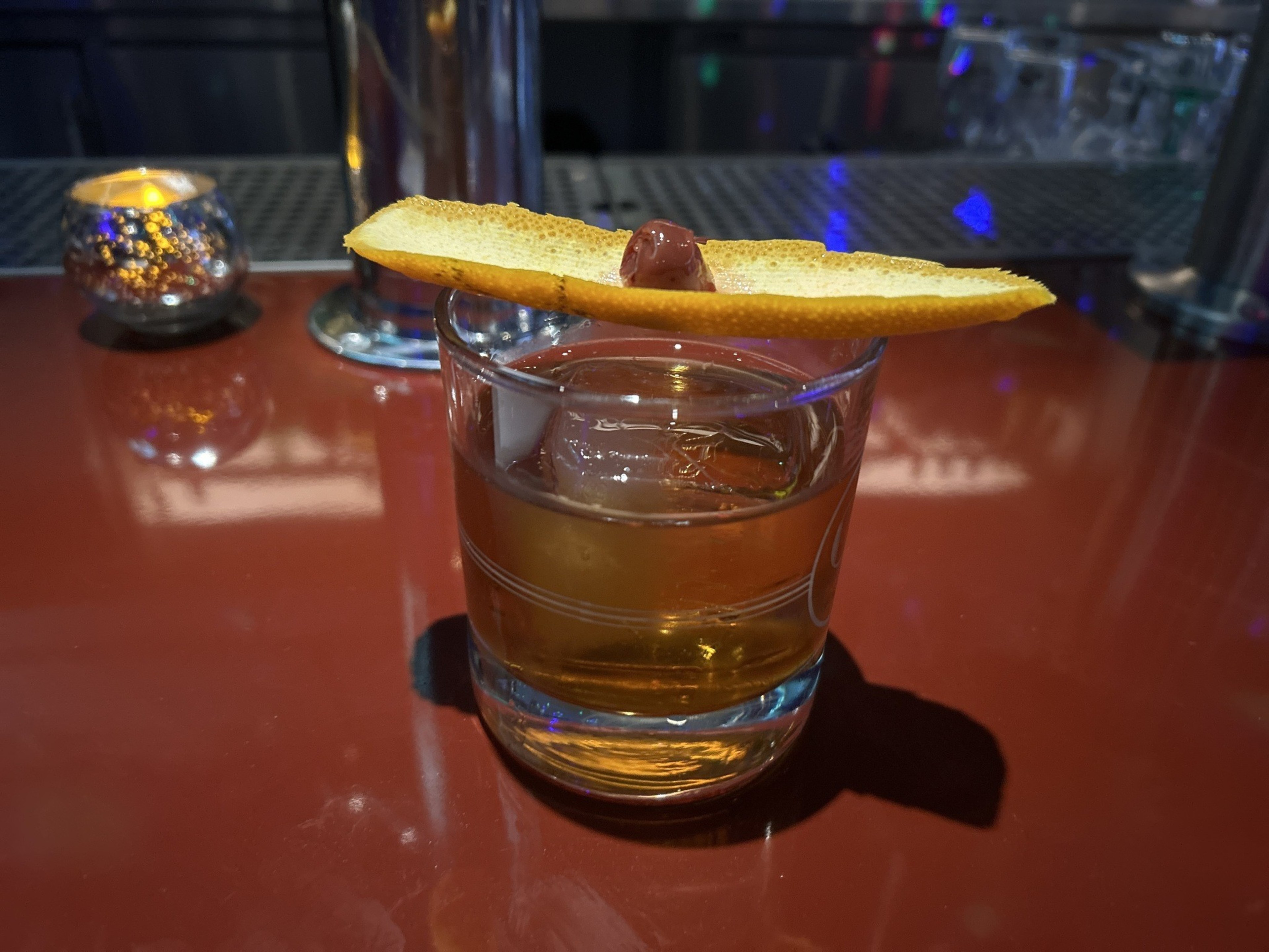A brown cocktail on the rocks garnished with an orange peel sits on a red bar.