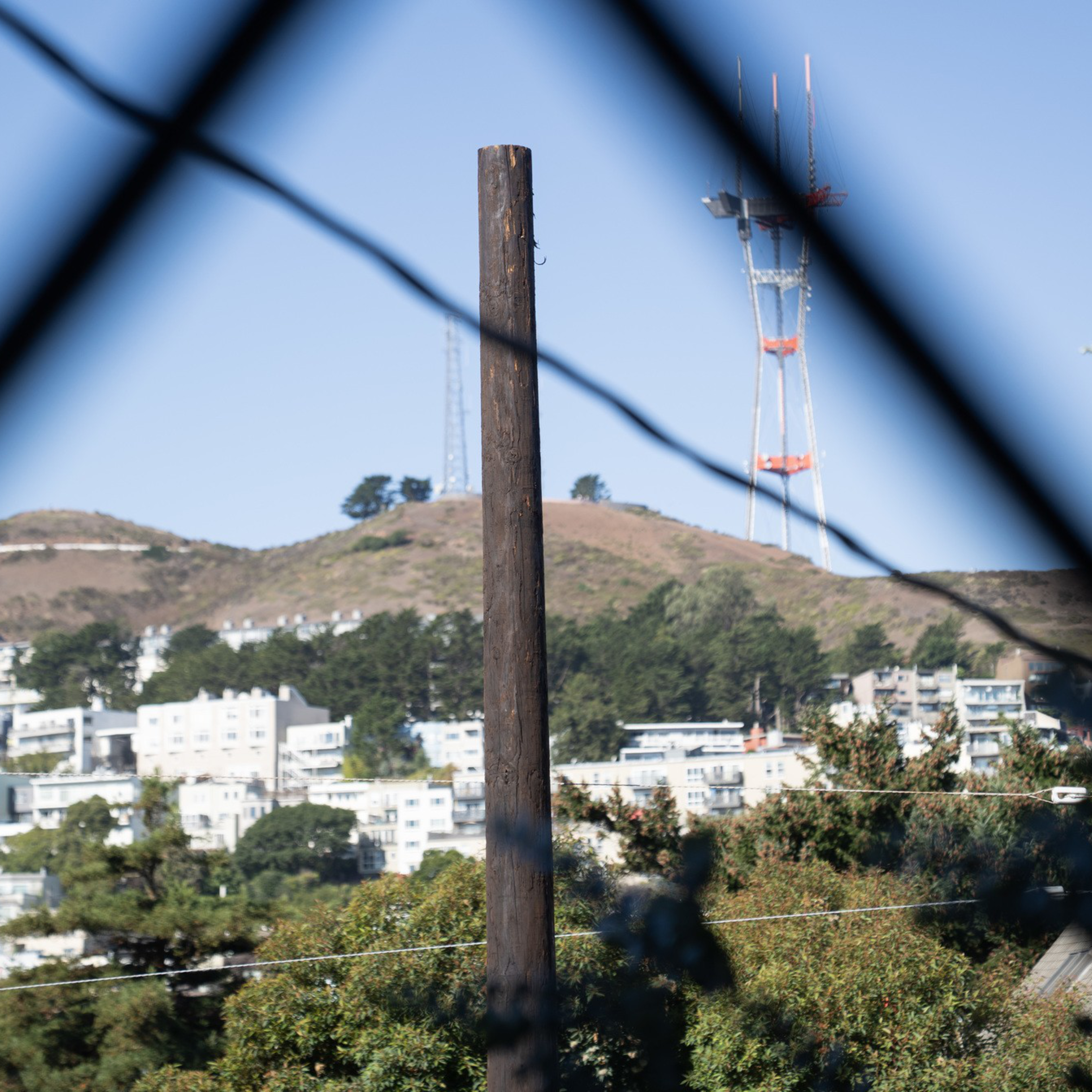 A tall communication tower stands on a hill behind a residential area, framed by a wooden pole and blurred fence wires in the foreground.