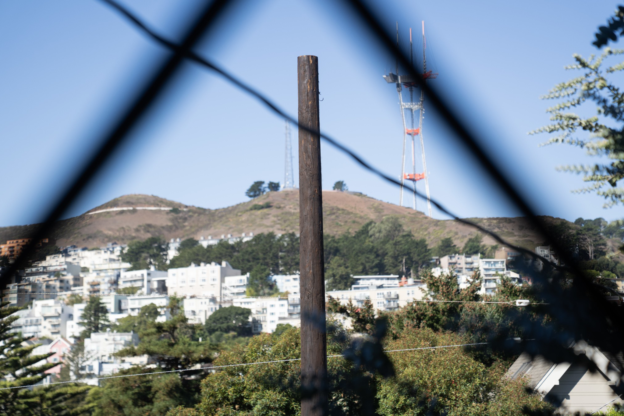 A tall communication tower stands on a hill behind a residential area, framed by a wooden pole and blurred fence wires in the foreground.
