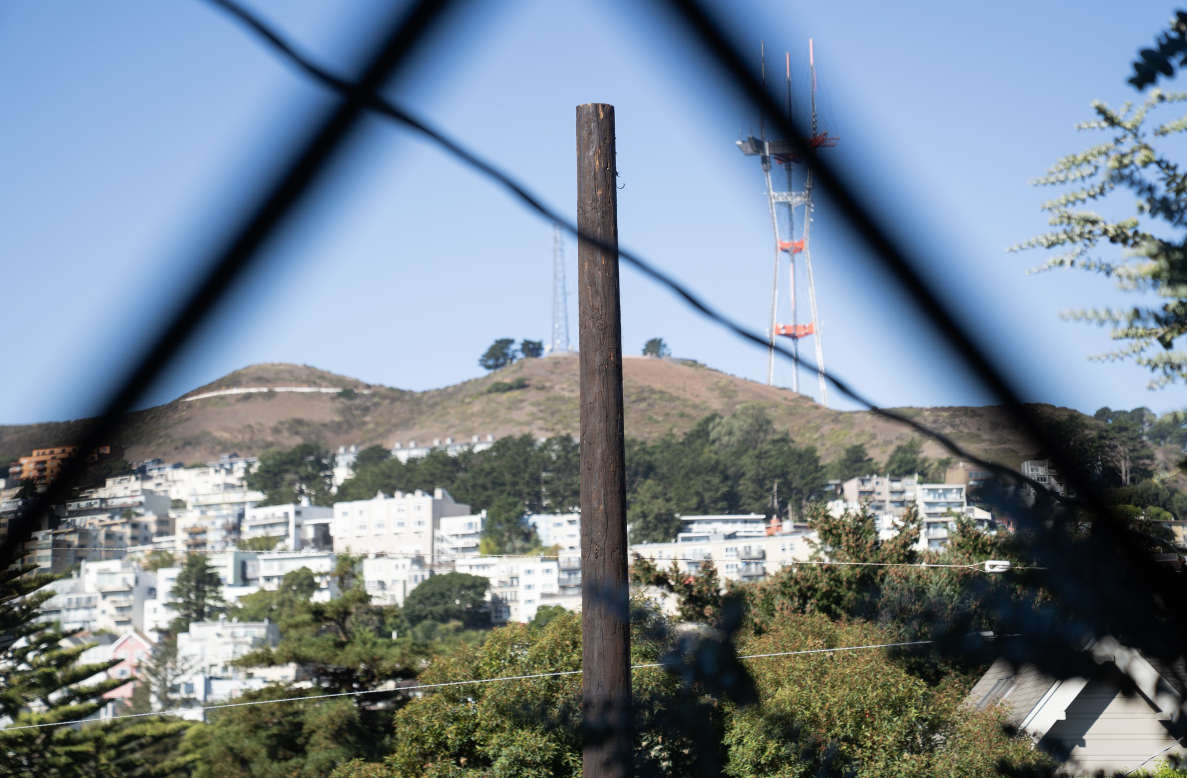 A tall communication tower stands on a hill behind a residential area, framed by a wooden pole and blurred fence wires in the foreground.