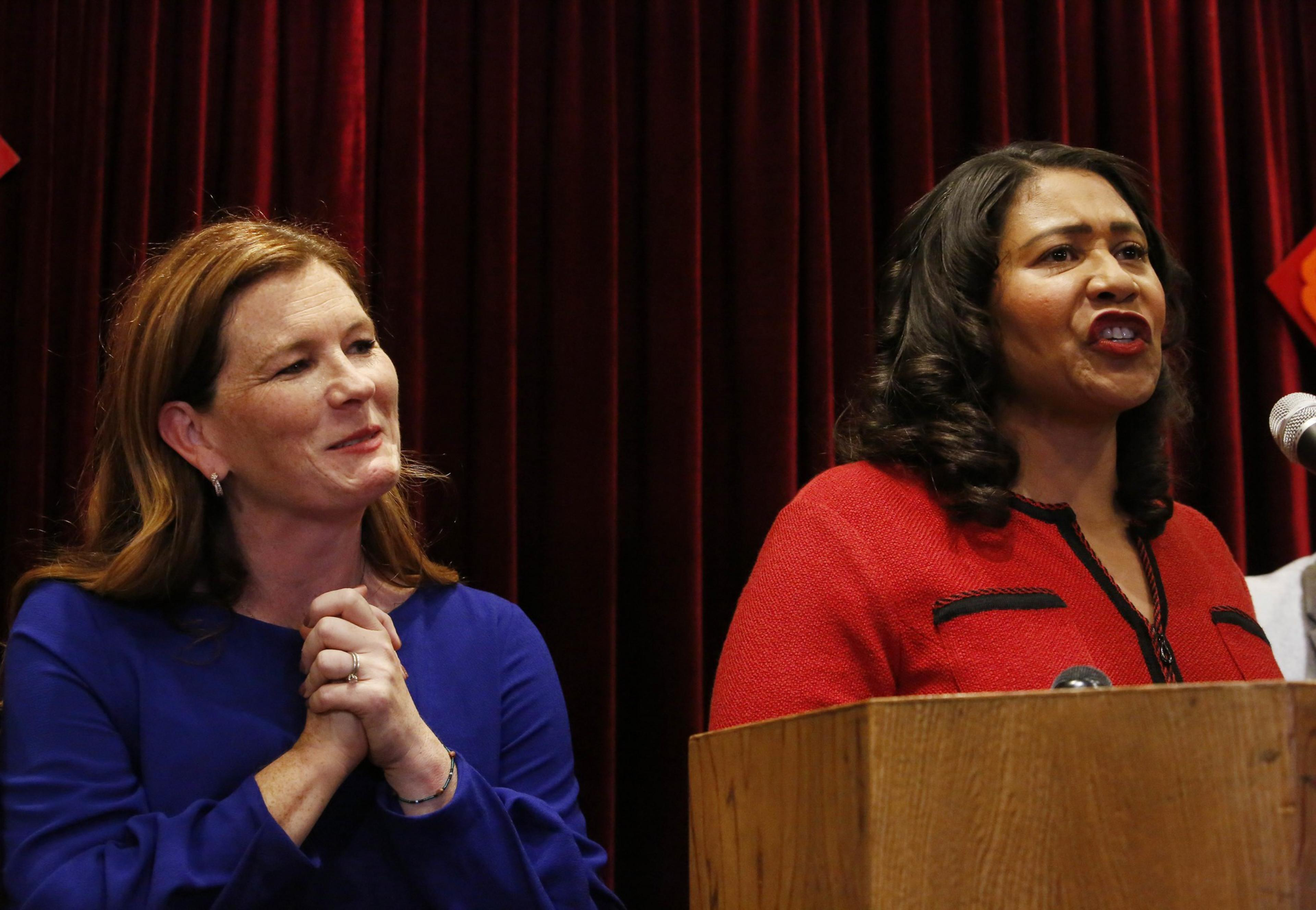 Two women are in front of a red curtain. One is speaking at a podium, wearing a red outfit, and the other stands next to her in blue, smiling and clasping her hands.