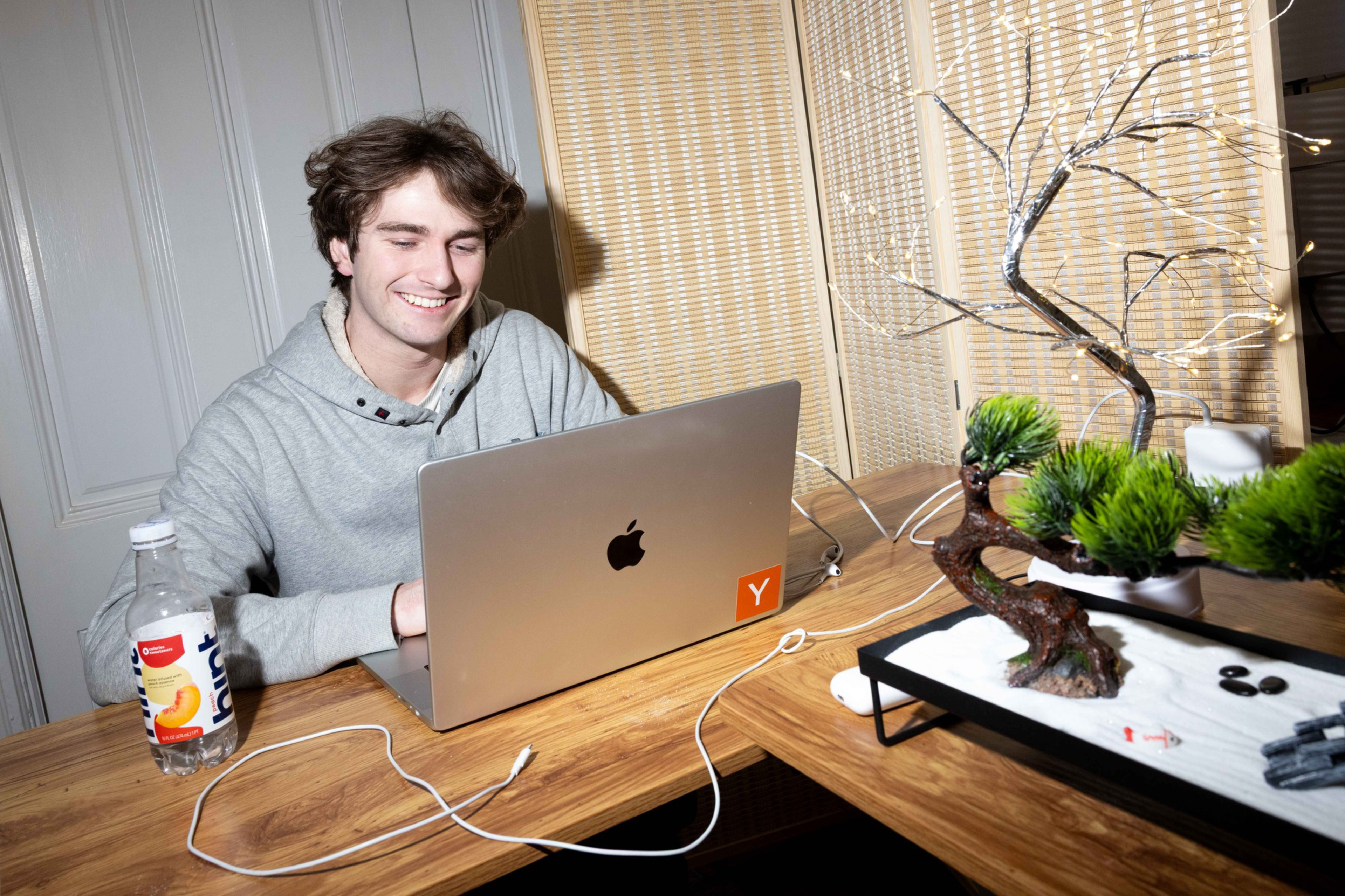 A person in a gray hoodie smiles while using a laptop on a wooden table. Nearby, thereâ€™s a bottle of water and a decorative bonsai tree with lights.