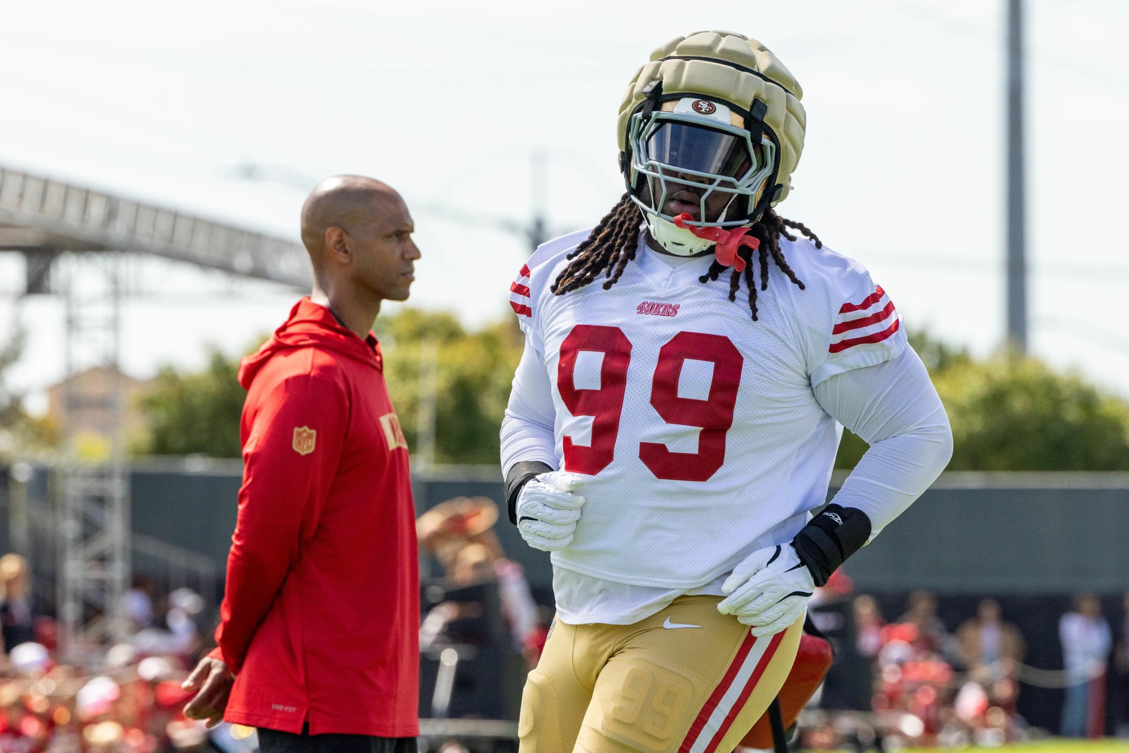 A football player in full gear and a helmet jogs during practice, wearing a white jersey with the number 99. A man in a red hoodie observes nearby.