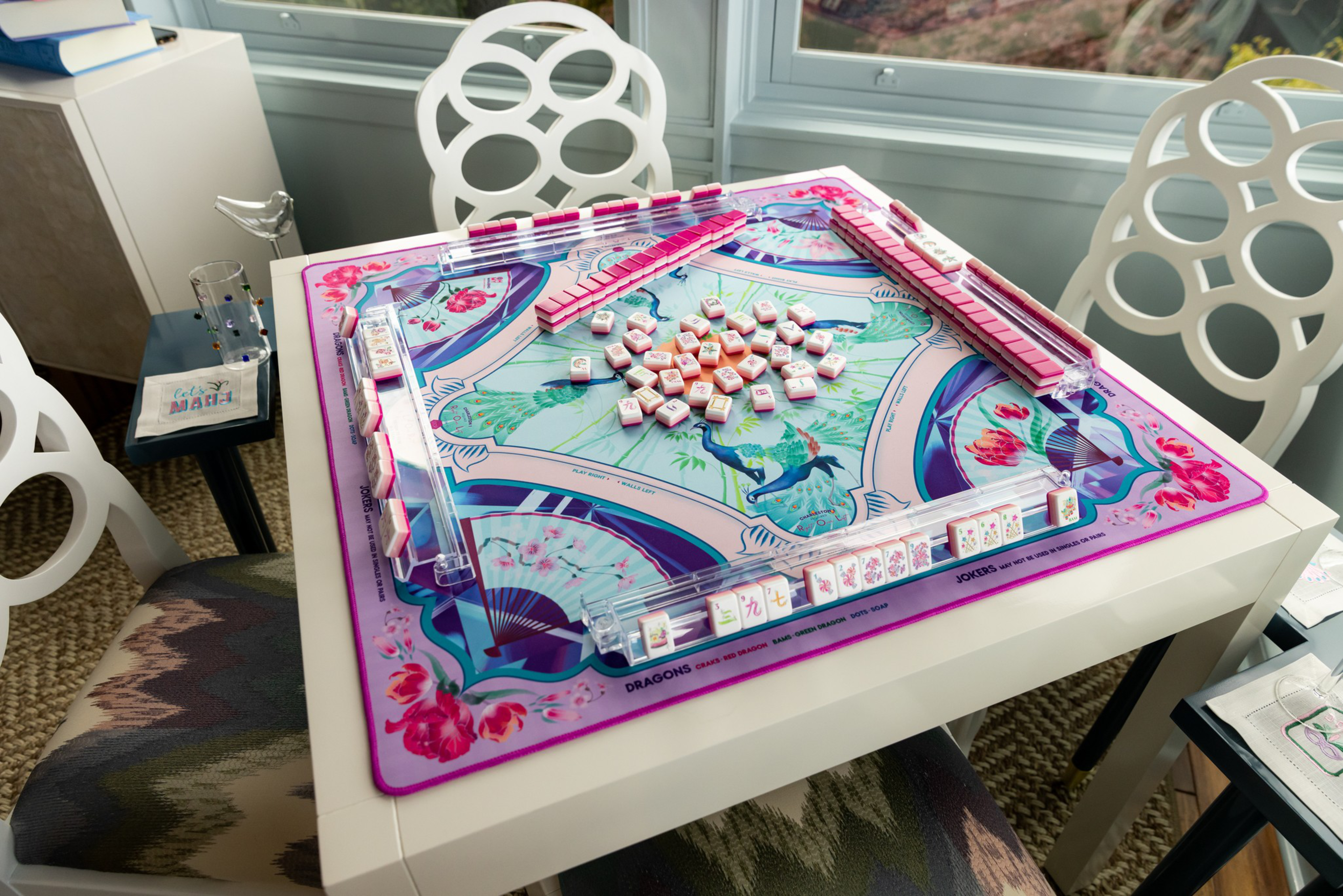 A colorful Mahjong game is set up on a white table with pink tiles and a floral-themed mat, surrounded by modern white chairs and a nearby table.