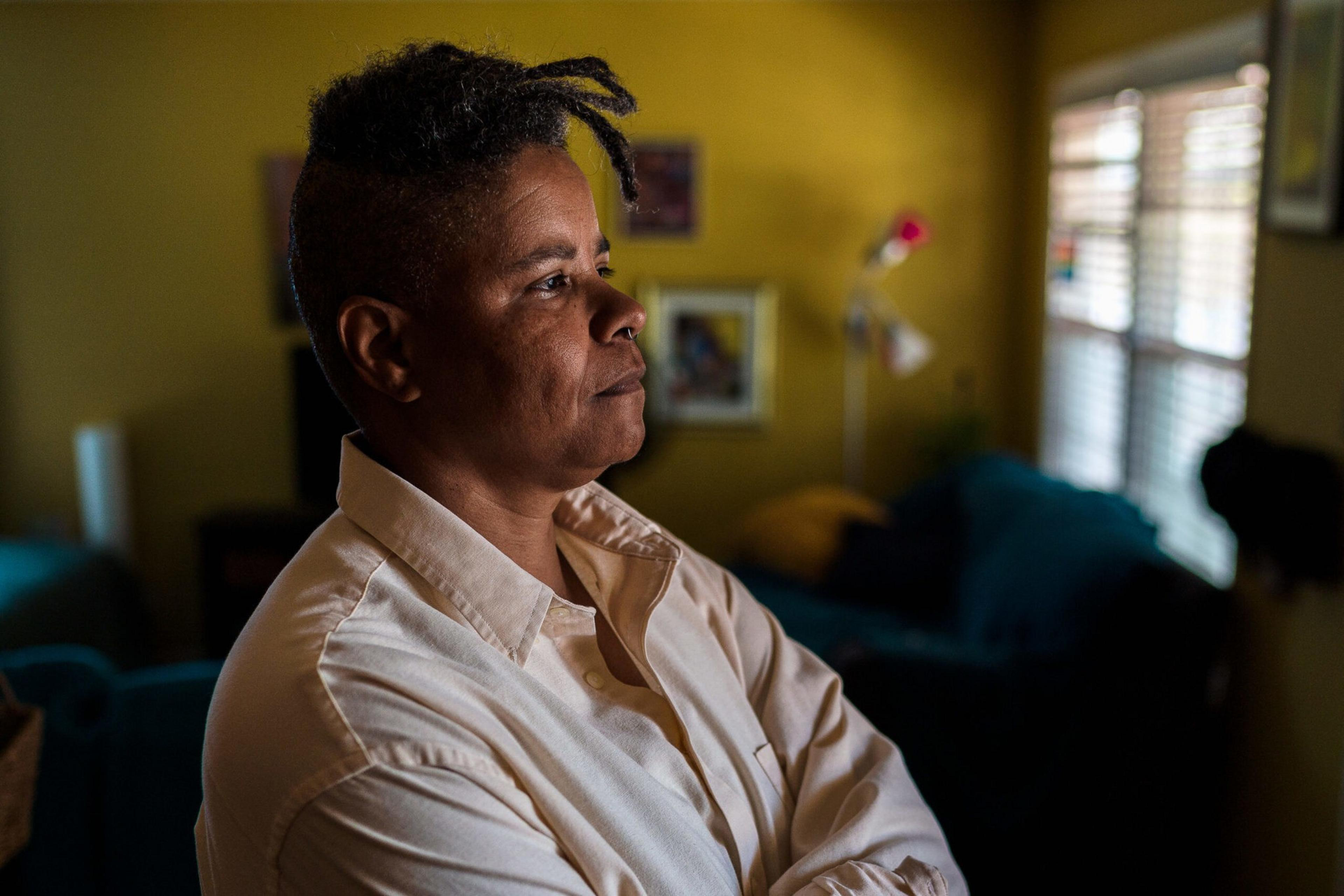 A person with short dreadlocks and wearing a white button-up shirt stands with arms crossed, looking thoughtfully out a window in a dim room.