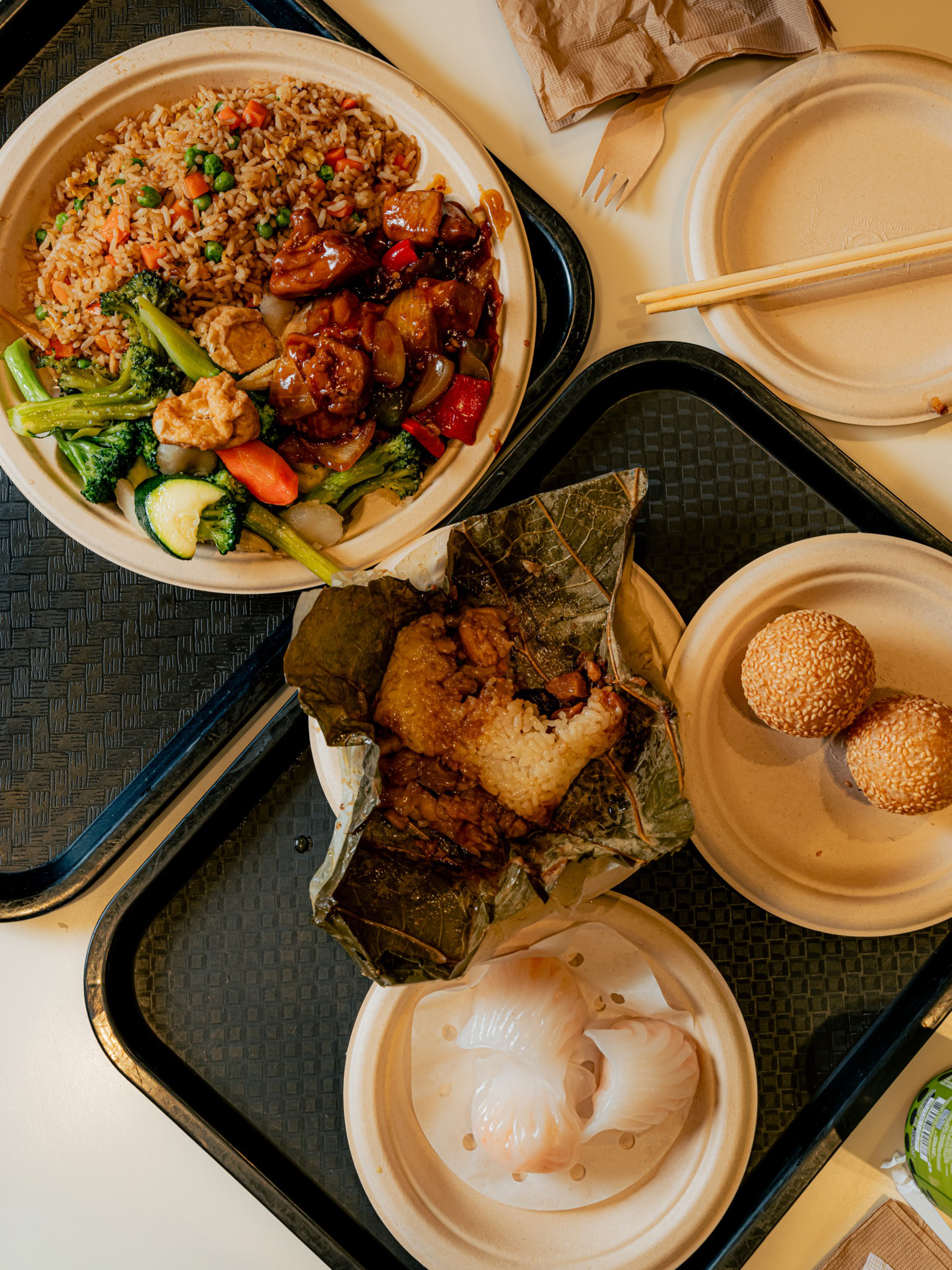 A meal with fried rice, stir-fried vegetables and meat, sticky rice wrapped in a leaf, two shrimp dumplings, and two sesame balls on separate plates.