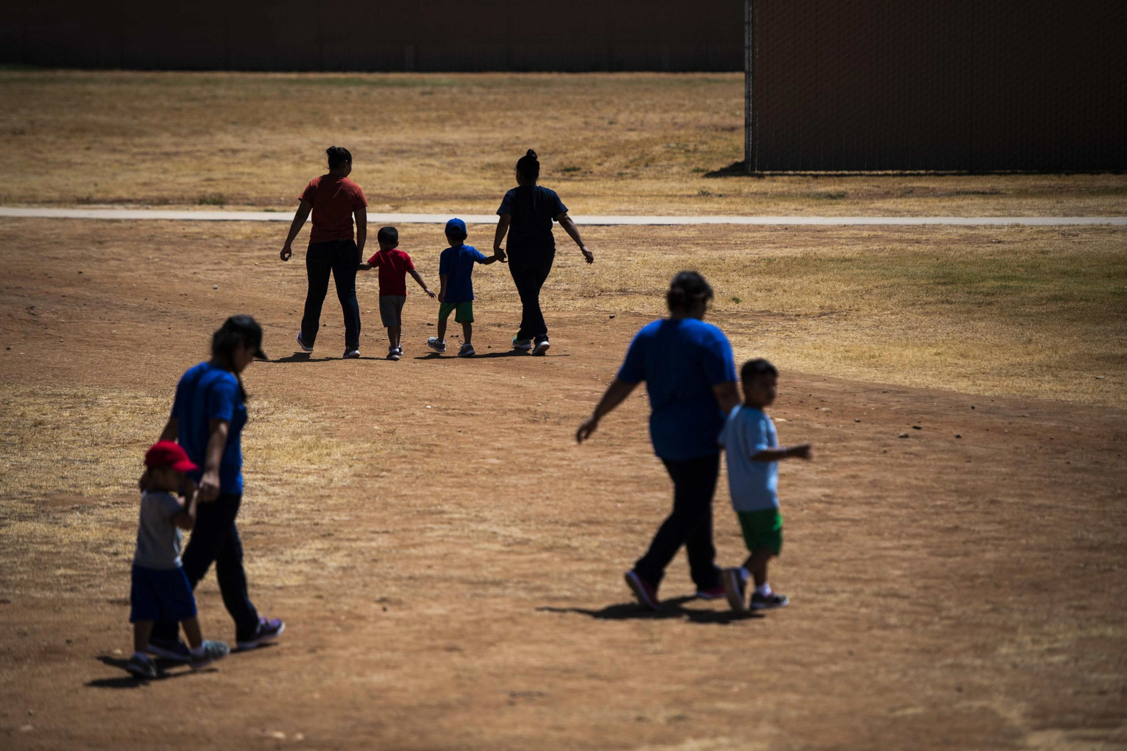 Several adults and children walk across a dry, brown field, holding hands. The background features a building, and the scene is sunny with visible shadows.