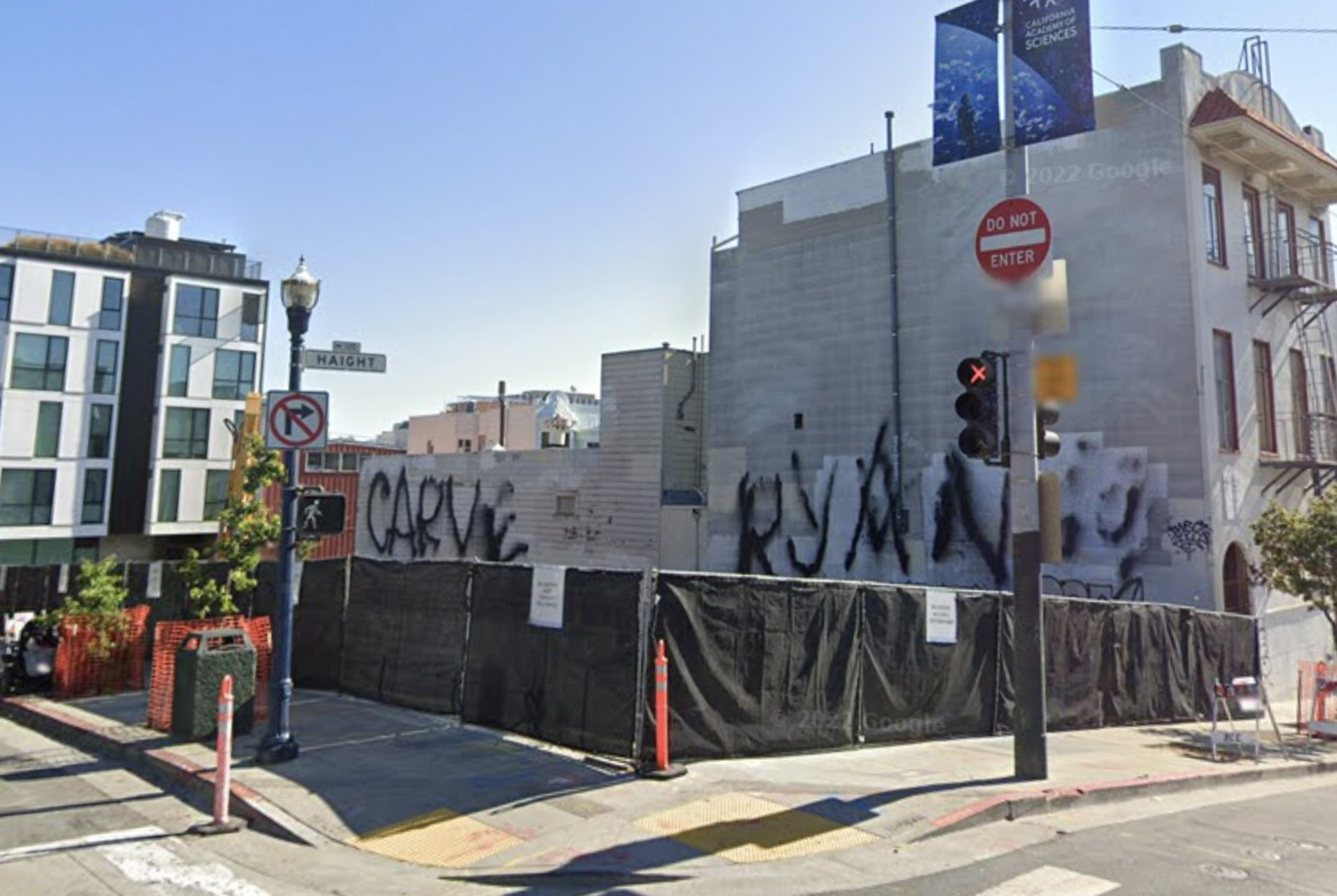 A street corner with buildings covered in large graffiti, black fencing around the site, street signs for Haight and no left turn visible.