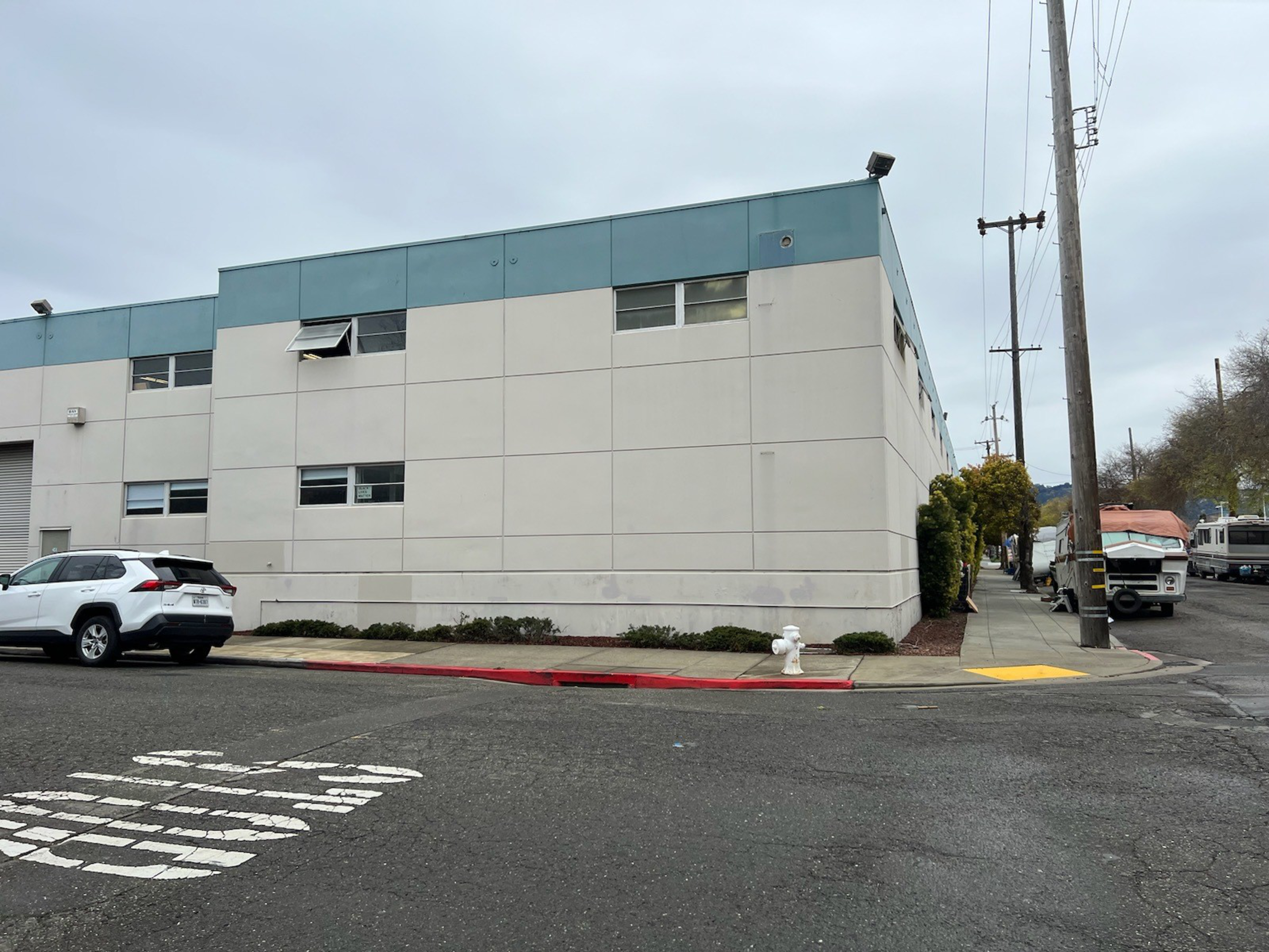 A white and teal industrial building sits at a street corner with a white SUV parked nearby, a fire hydrant, and power lines lining the sidewalk.
