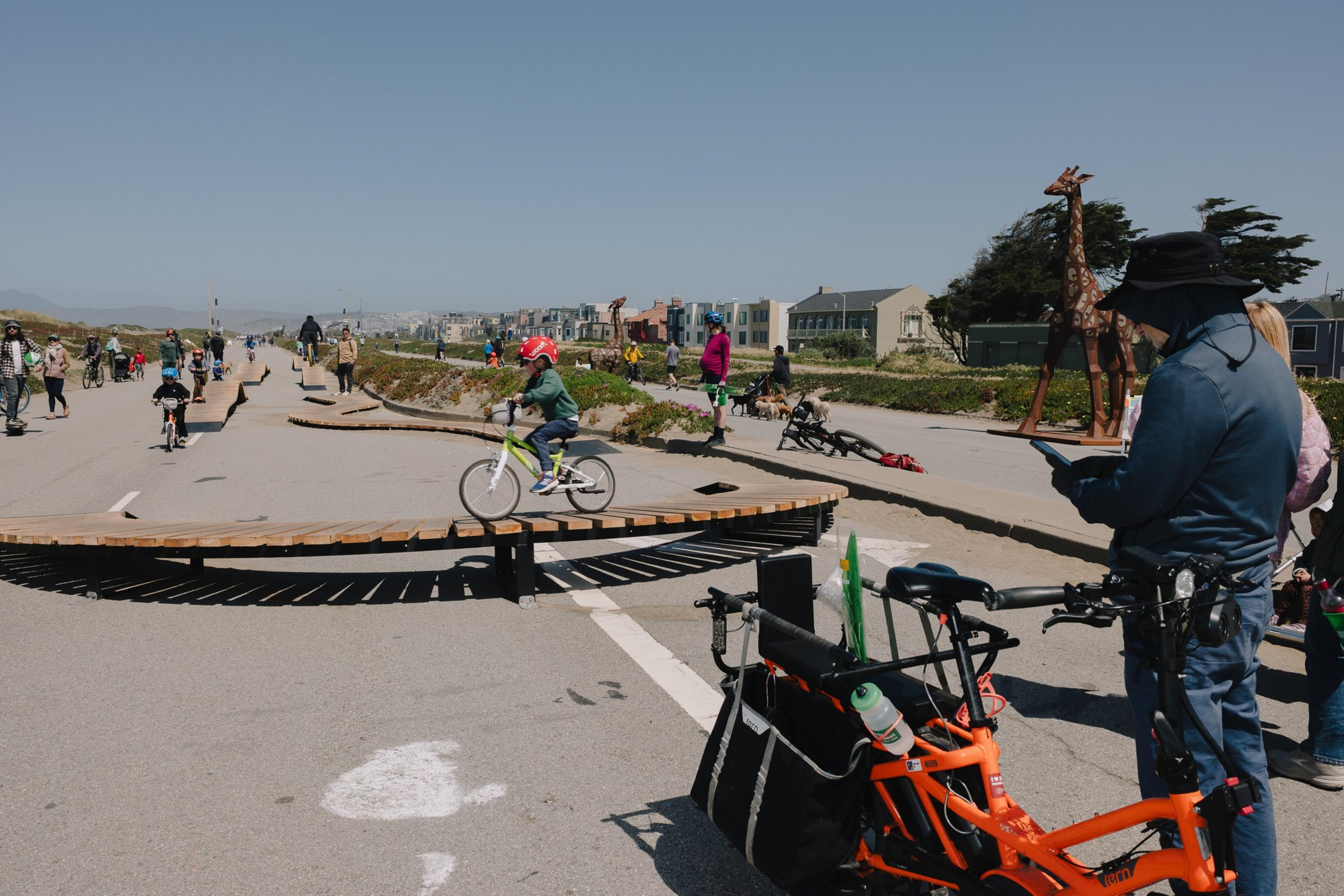 People are enjoying a sunny day outdoors. Kids on bikes ride over a wooden ramp, while adults watch nearby. A wooden giraffe sculpture stands in the background.