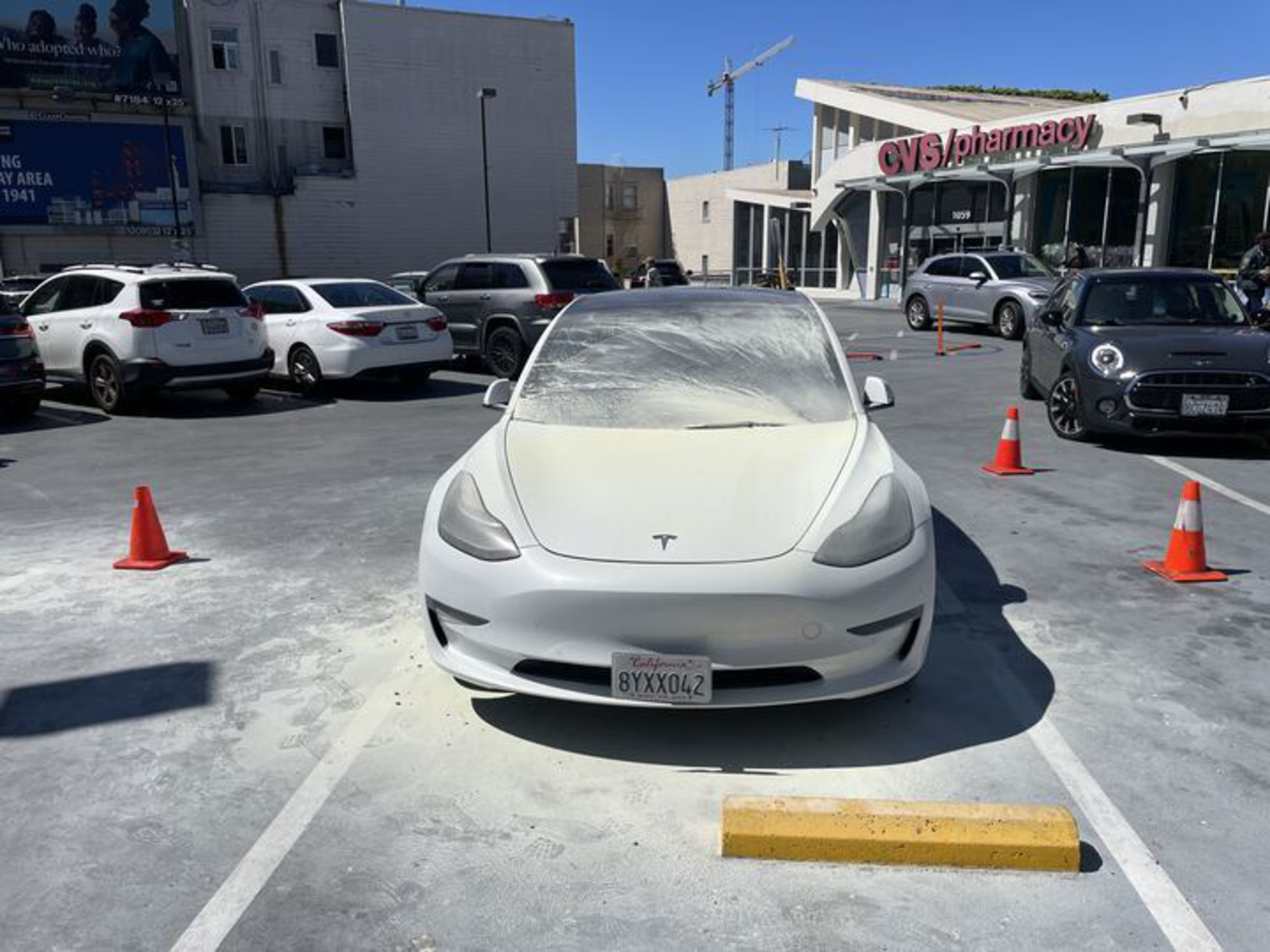 A white Tesla with a dusty windshield is parked in a spot near a CVS pharmacy, surrounded by orange traffic cones and other cars.