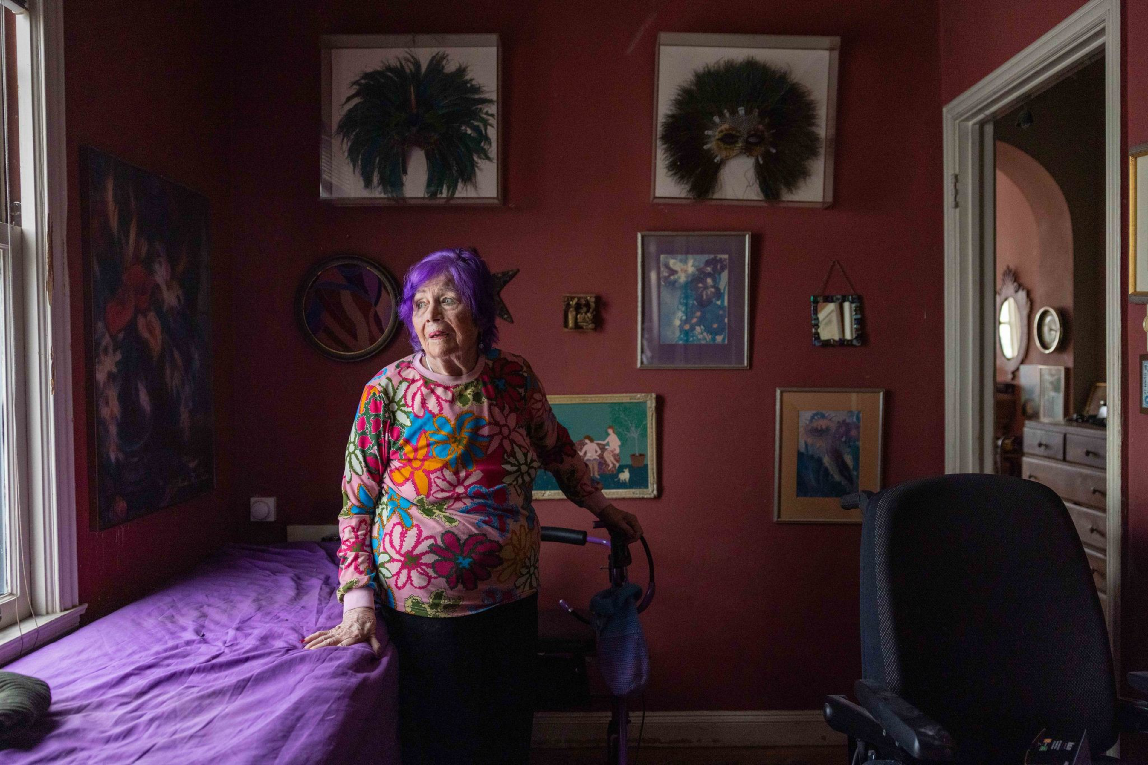 An elderly woman with purple hair and a colorful floral top stands in a dimly lit room with deep red walls, surrounded by framed art and a walker.