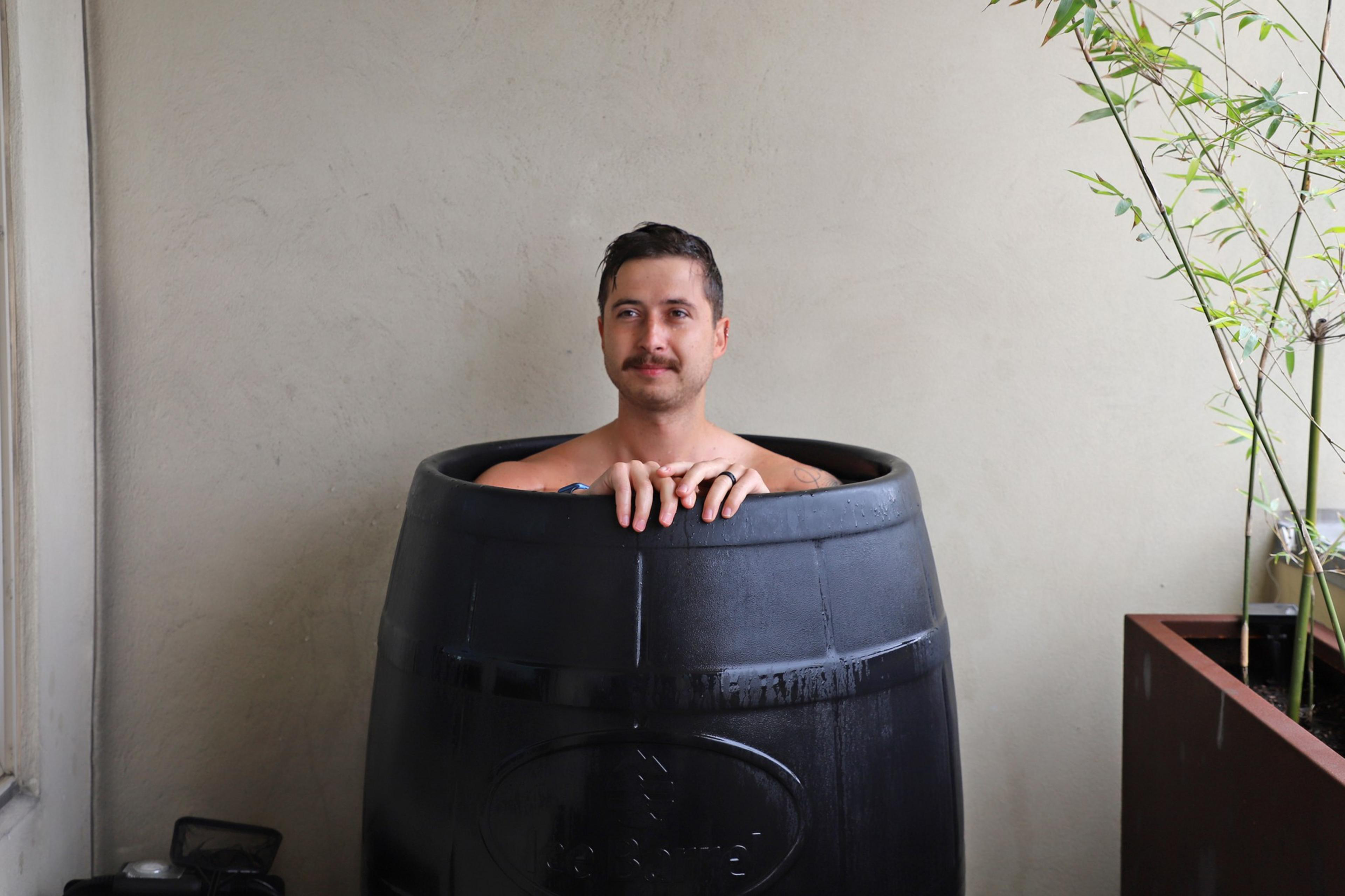 A man with short dark hair and a mustache sits inside a large black barrel, with only his shoulders and hands visible above the rim.