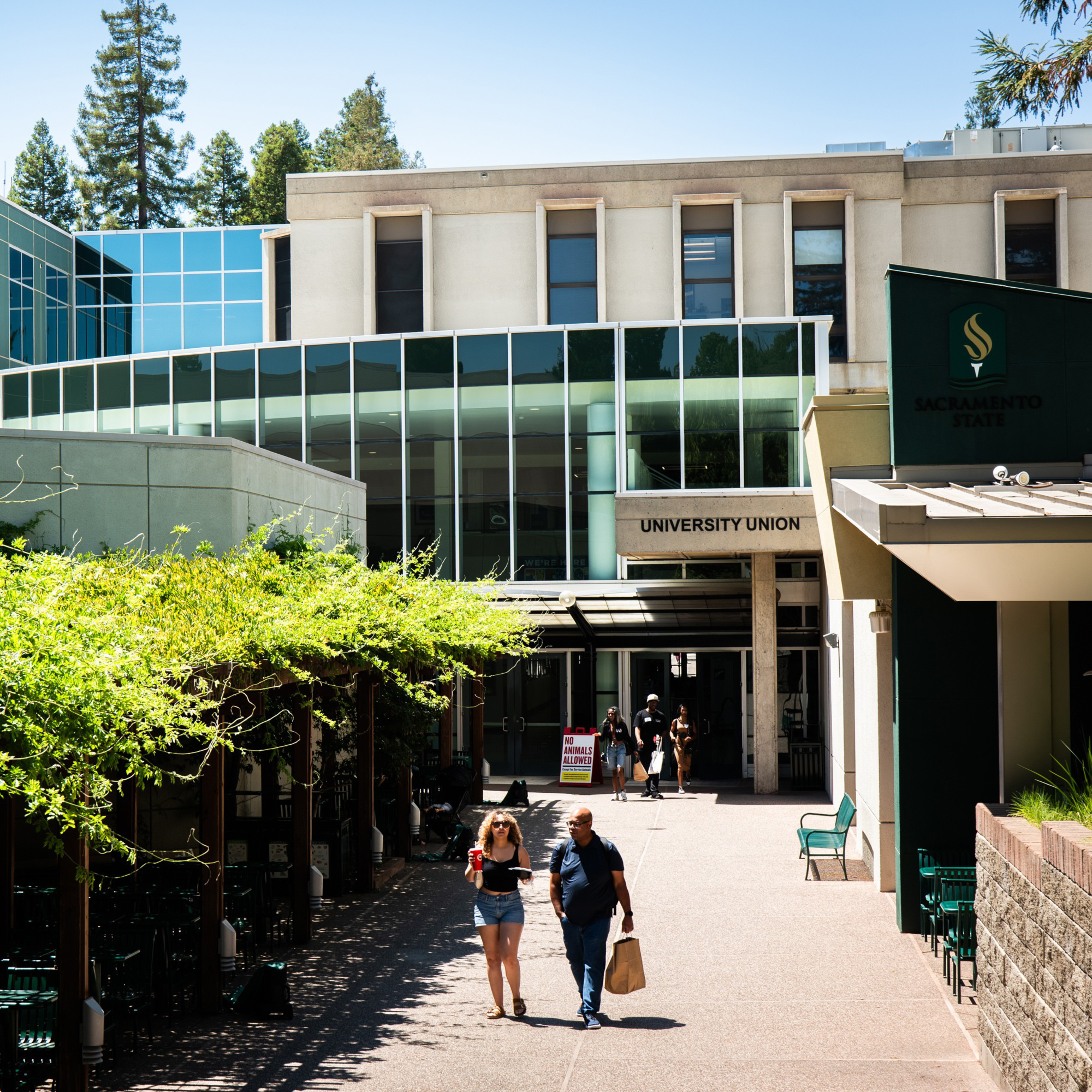 The image shows the entrance to the University Union building at Sacramento State. People are walking on a pathway surrounded by greenery and modern architecture.