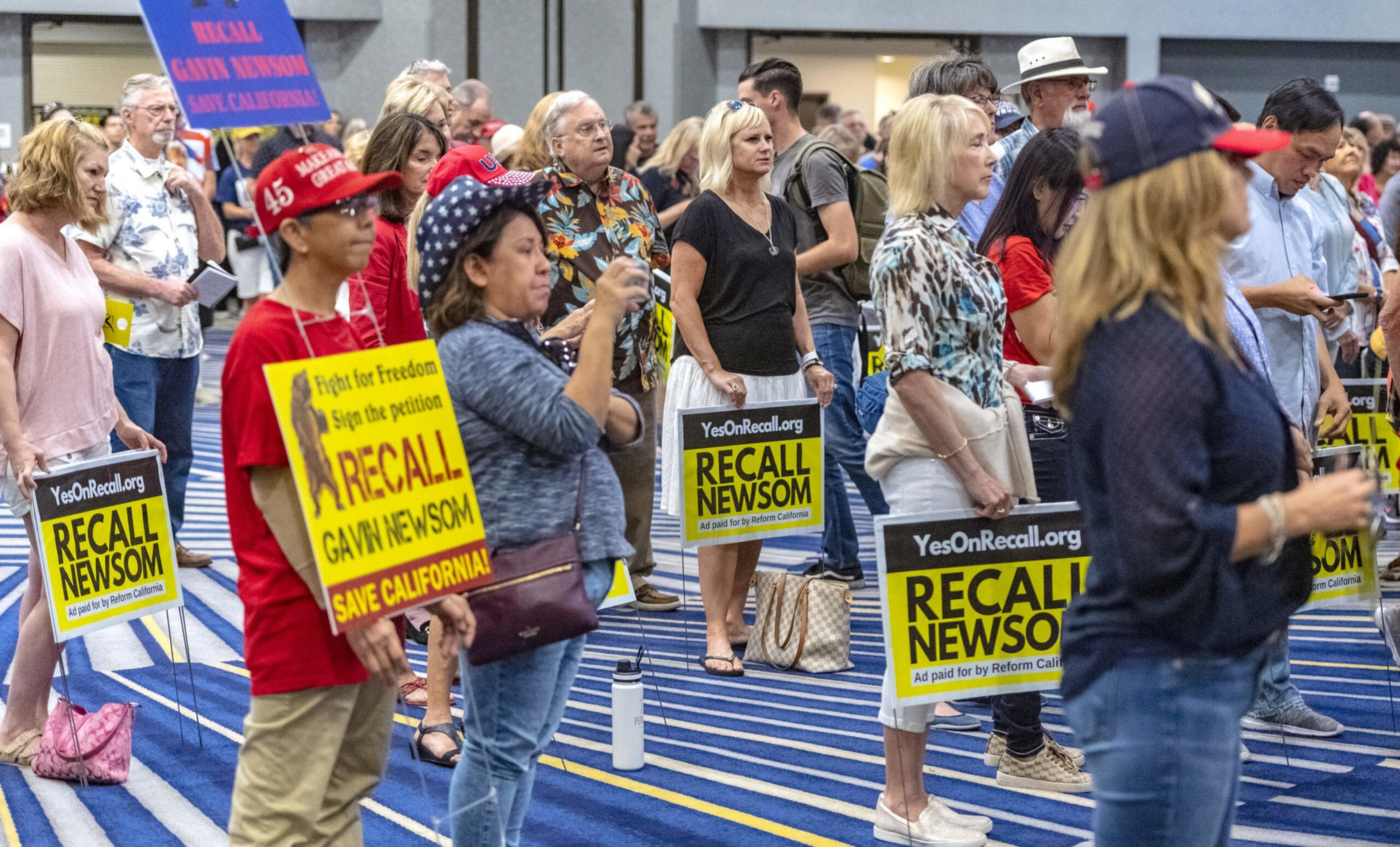 A diverse group of people holds yellow and black signs reading "Recall Newsom," gathered indoors on a blue-striped carpet.