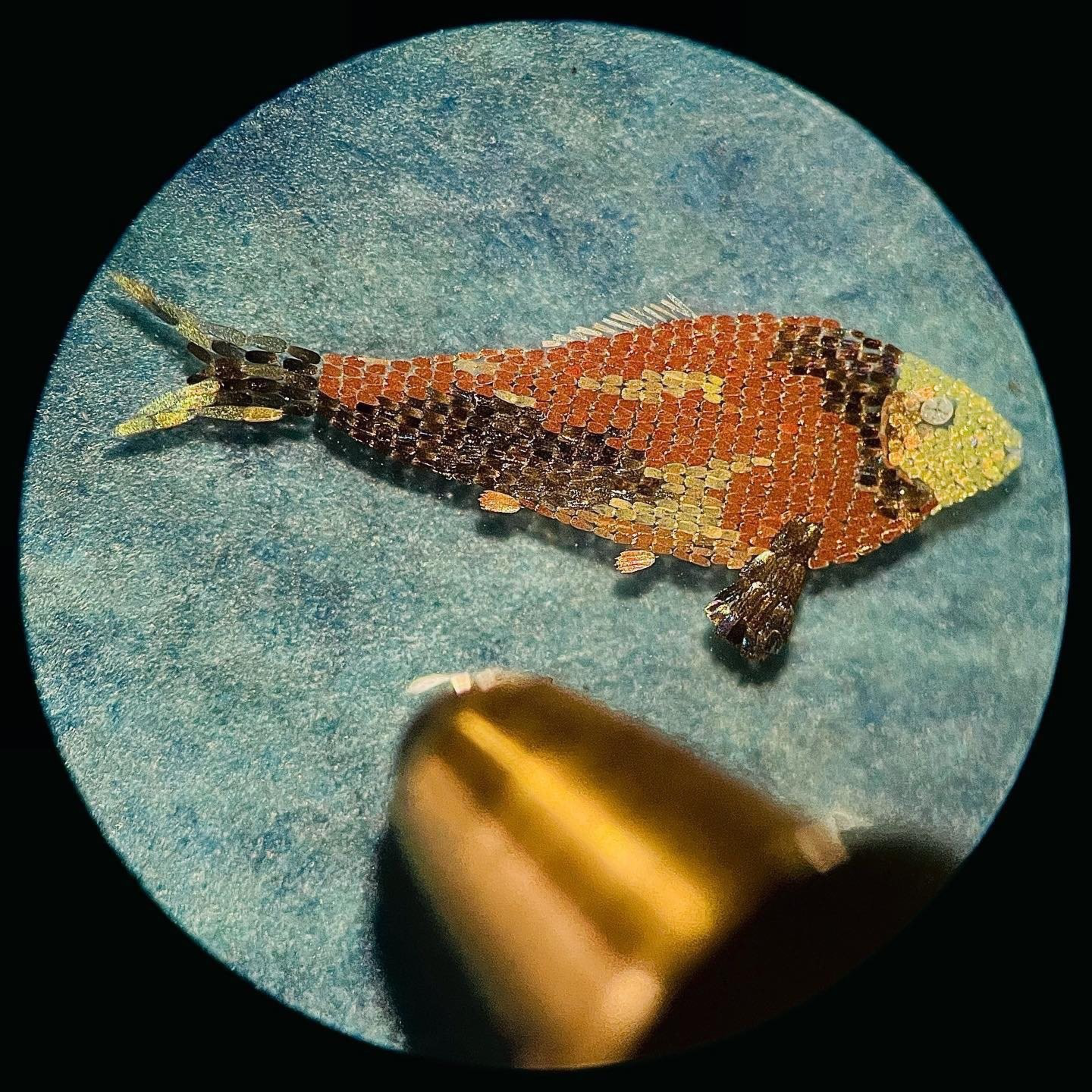 A tiny, detailed fish mosaic made of multicolored tiles sits on a blue background, with a pen tip nearby for scale, showing its minuscule size.
