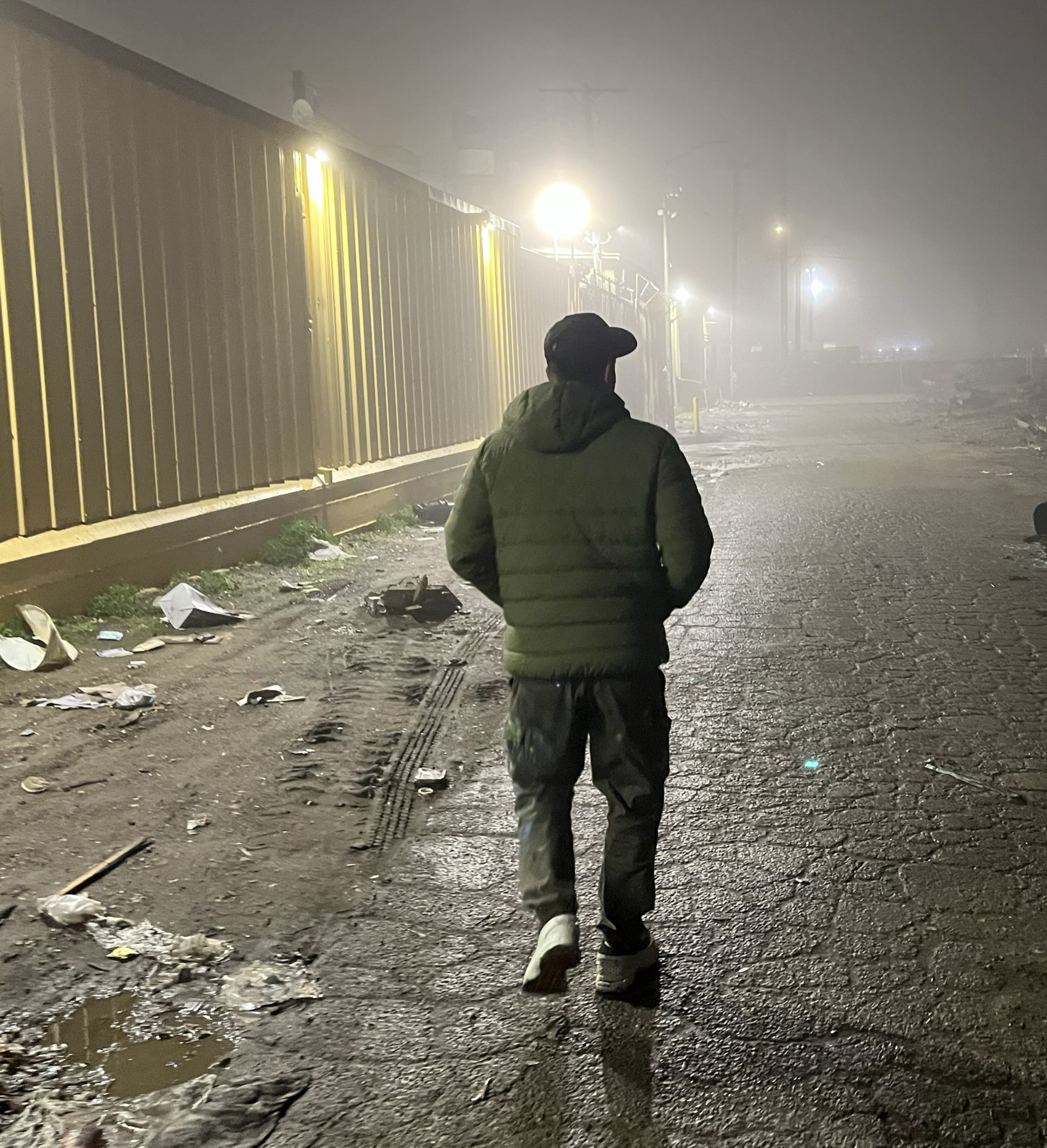 A person in a hooded jacket and cap walks on a dimly lit, foggy street. Trash is scattered on the ground near a long building with bright overhead lights.