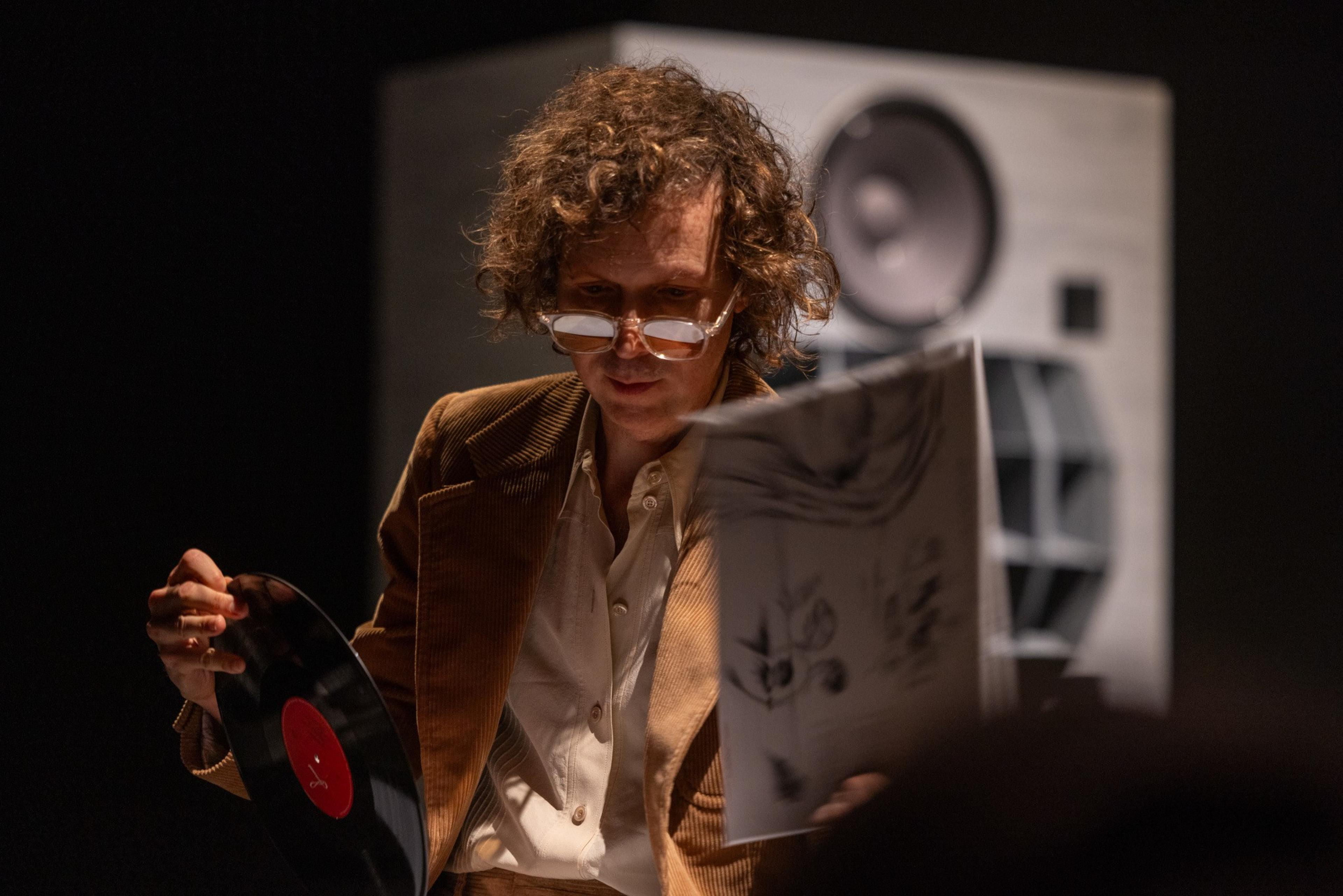 A person with curly hair, wearing glasses and a brown jacket, is examining a vinyl record and its cover. There is a large speaker in the background.