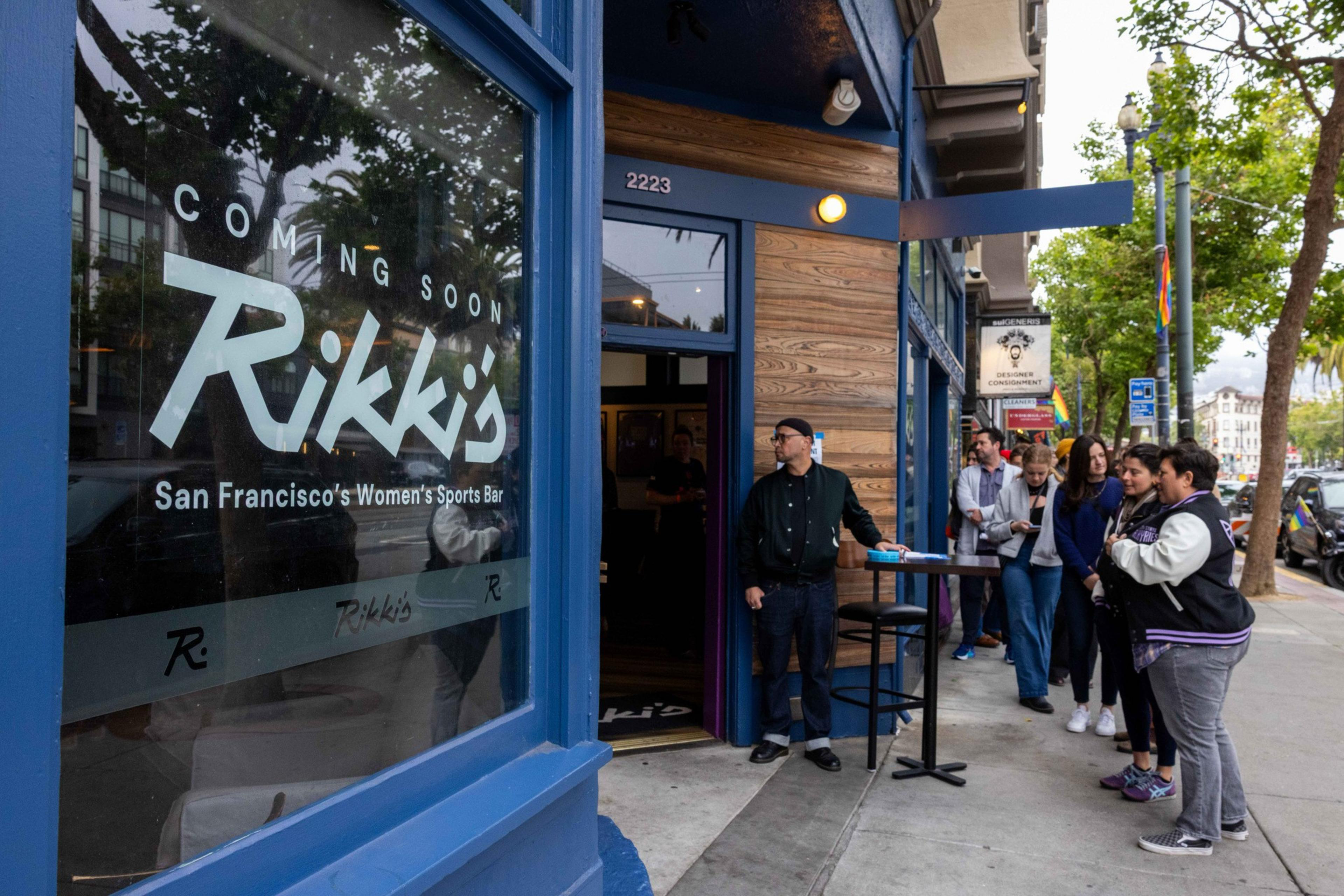 A group of people stands outside &quot;Rikki's,&quot; a soon-to-open women's sports bar in San Francisco. The entrance features a blue door and wood paneling.