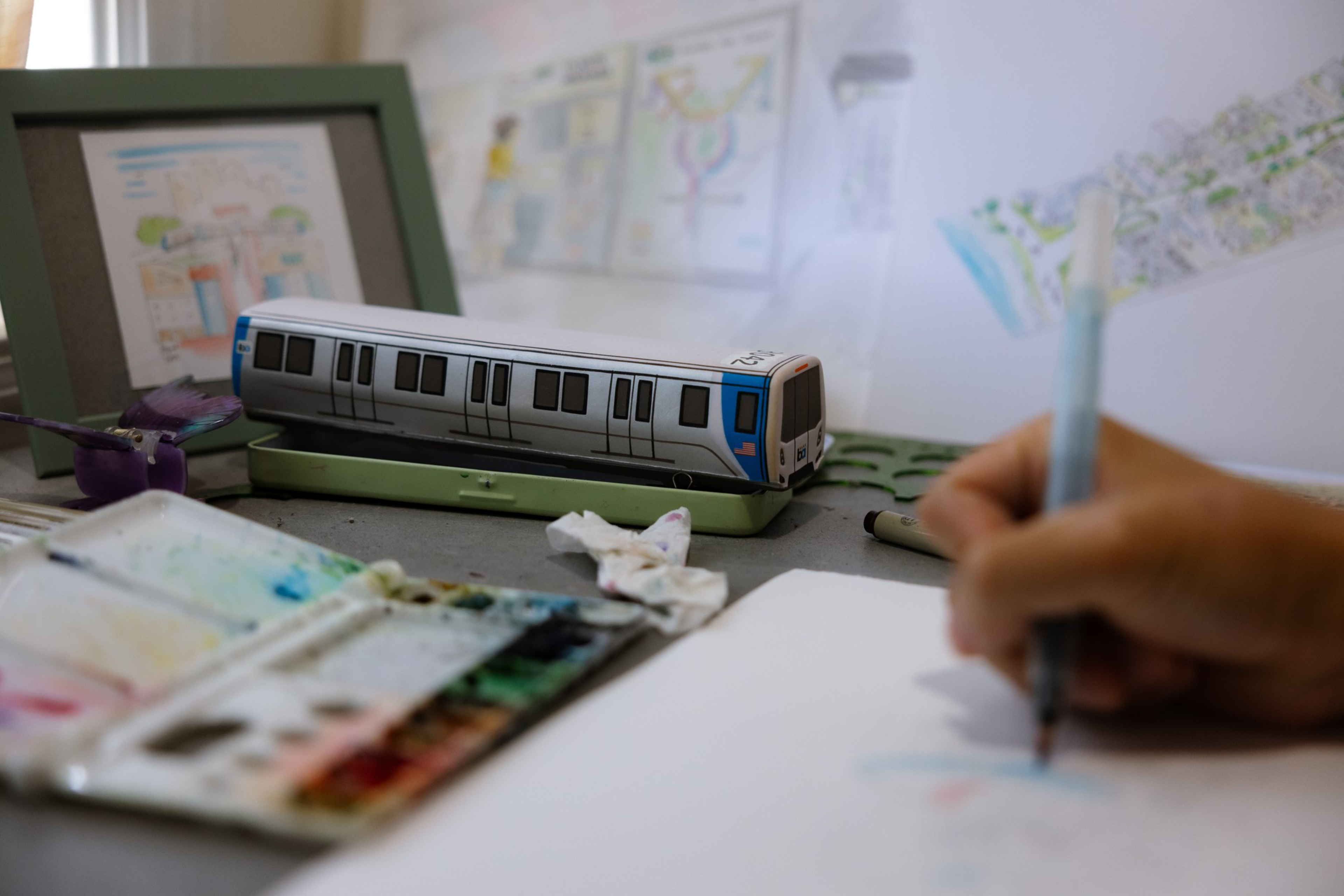 A hand is drawing on white paper with a marker next to a paint palette, a miniature train model, and sketches pinned on a wall.