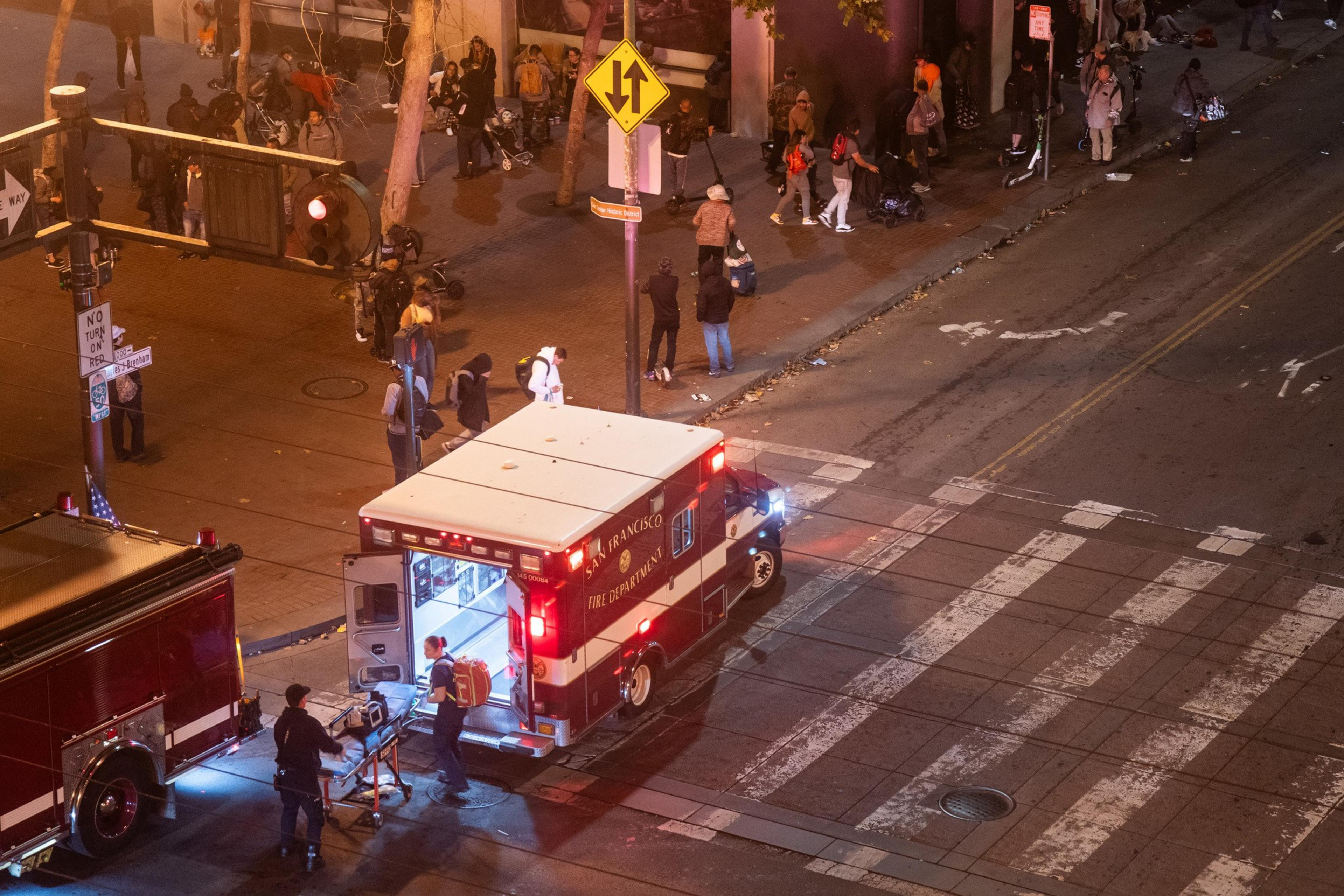A nighttime urban scene shows an ambulance and a fire truck at a crosswalk, with paramedics attending to someone. A crowd is gathered nearby on the sidewalk.