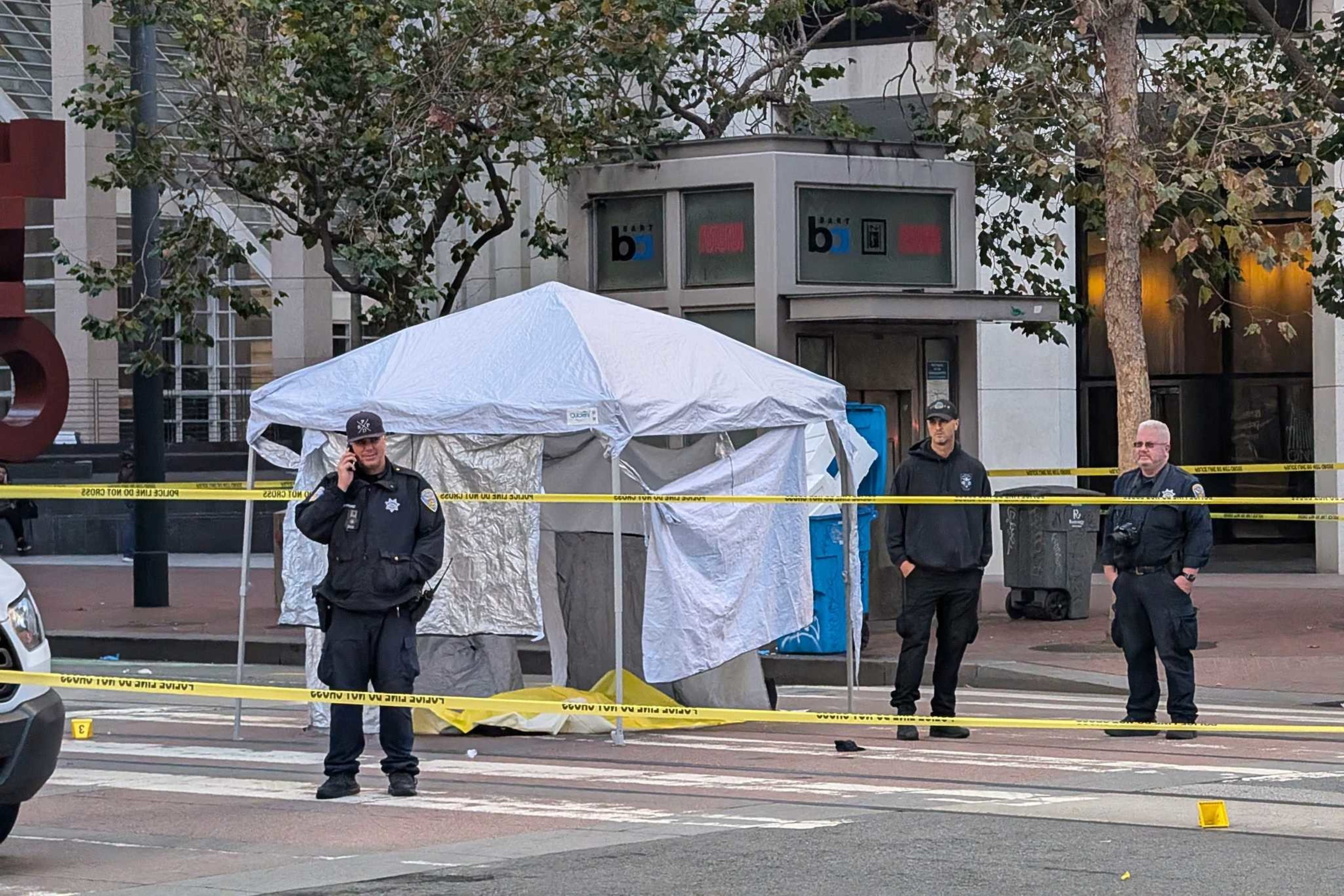 Police investigate a deadly stabbing in downtown San Francisco, near the Embarcadero station, on November 13, 2024.A street scene shows three police officers standing near a white tent, with yellow caution tape cordoning off the area. There are trees and a