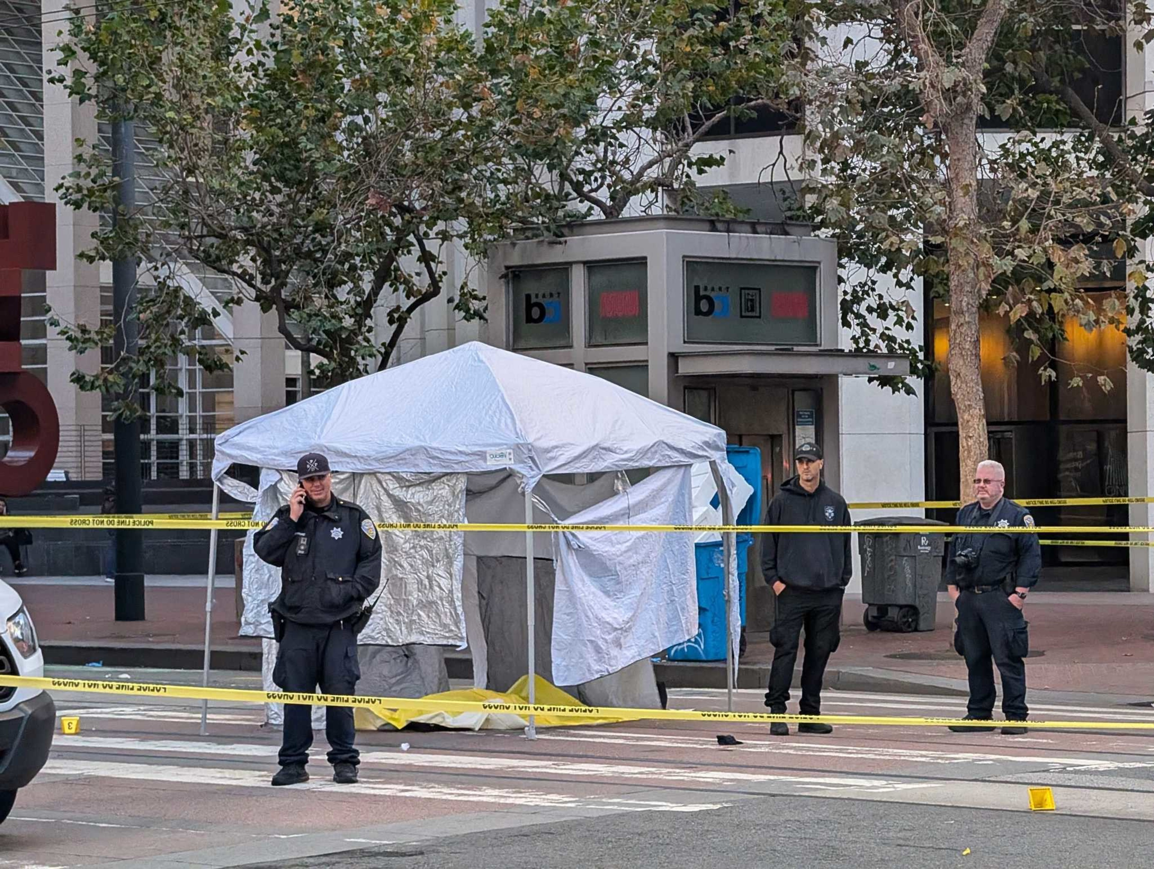Police investigate a deadly stabbing in downtown San Francisco, near the Embarcadero station, on November 13, 2024.A street scene shows three police officers standing near a white tent, with yellow caution tape cordoning off the area. There are trees and a
