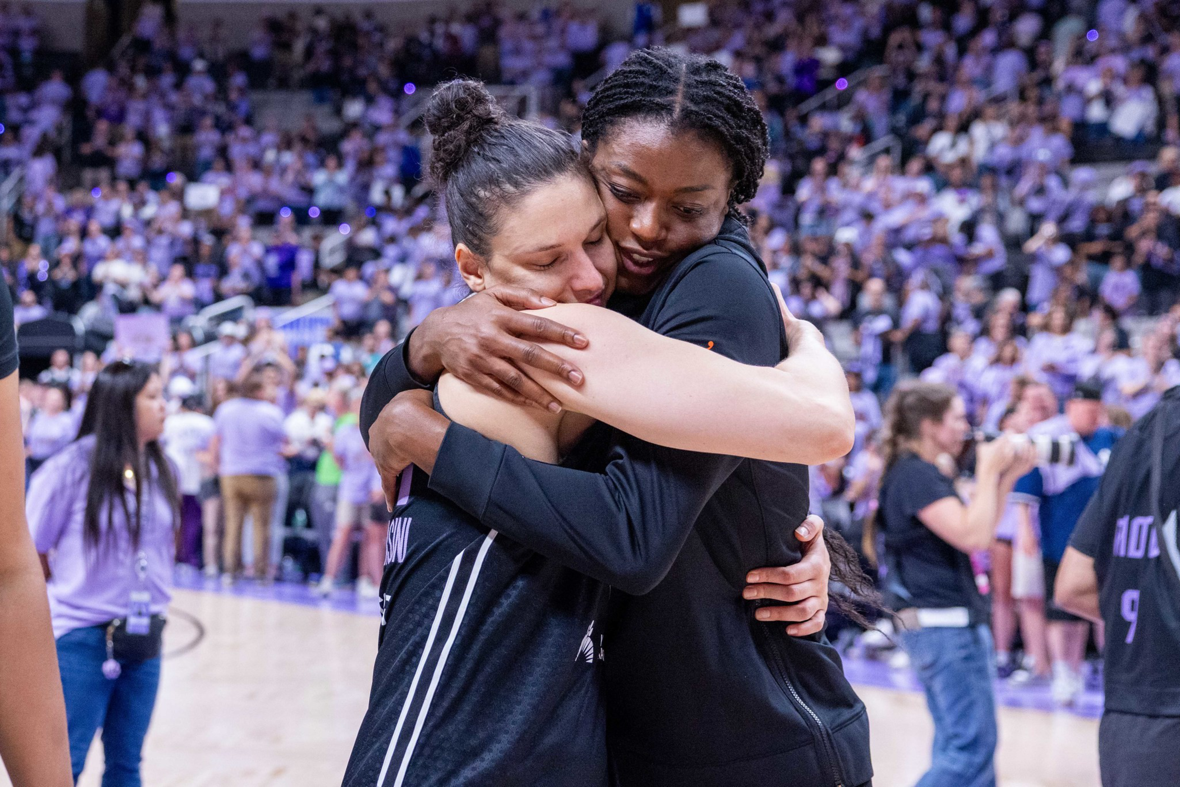 Two female basketball players tightly hug each other on the court, surrounded by a crowd of spectators.