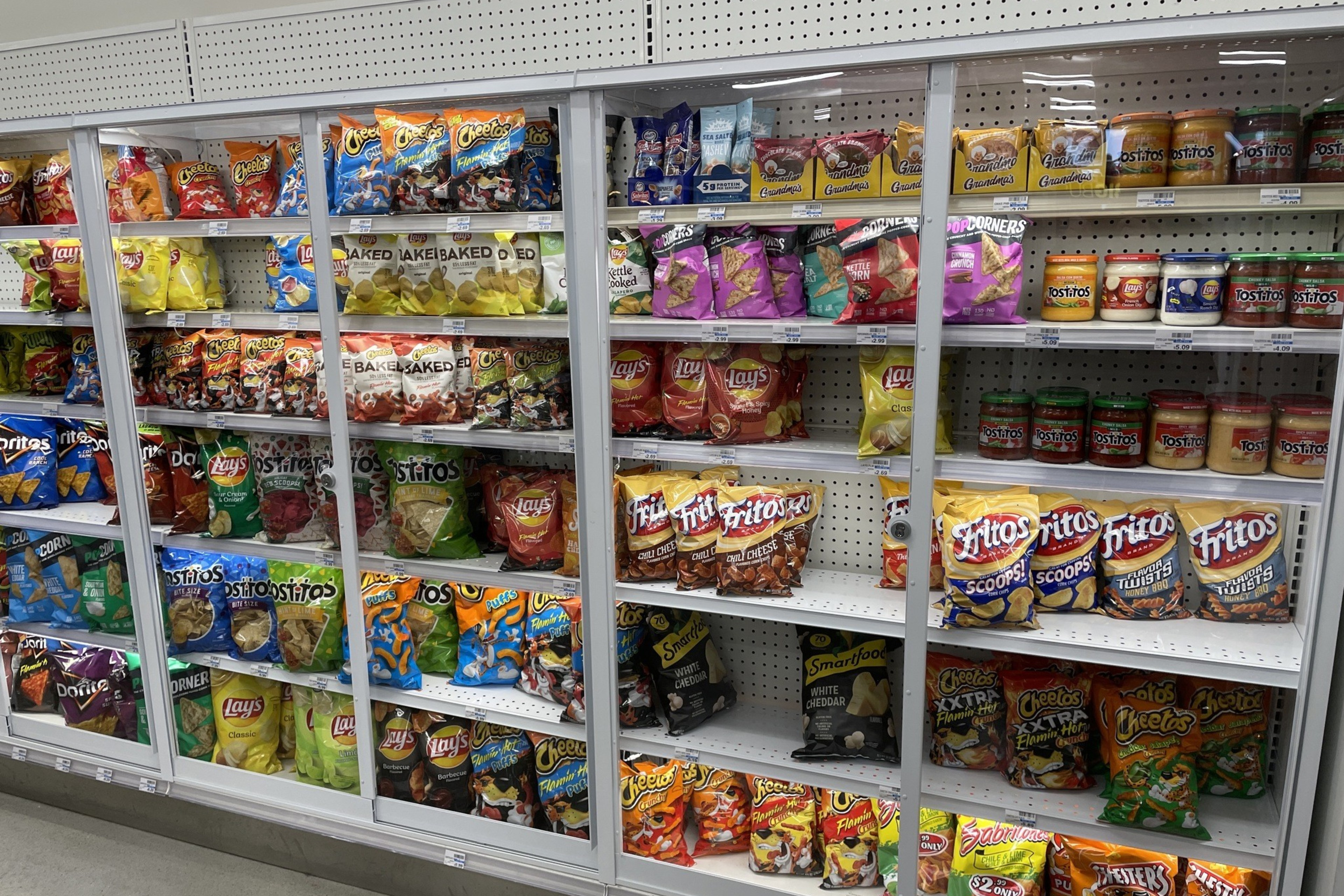 The image shows shelves in a store stocked with various chips and snacks, including brands like Cheetos, Fritos, Lays, Tostitos, and popcorn.