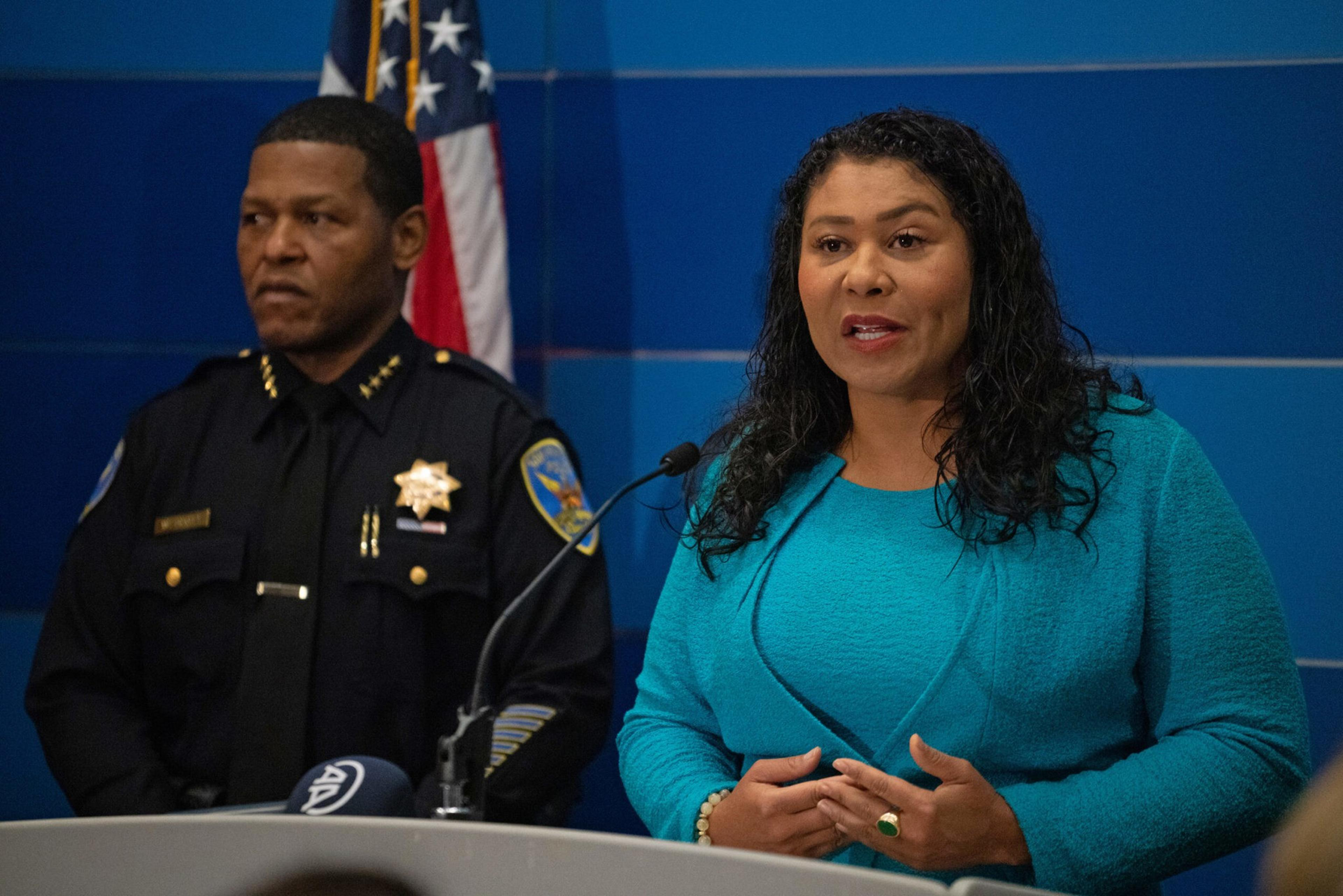 A woman in a turquoise outfit speaks at a podium with a microphone while a police officer in uniform stands behind her, with an American flag in the background.