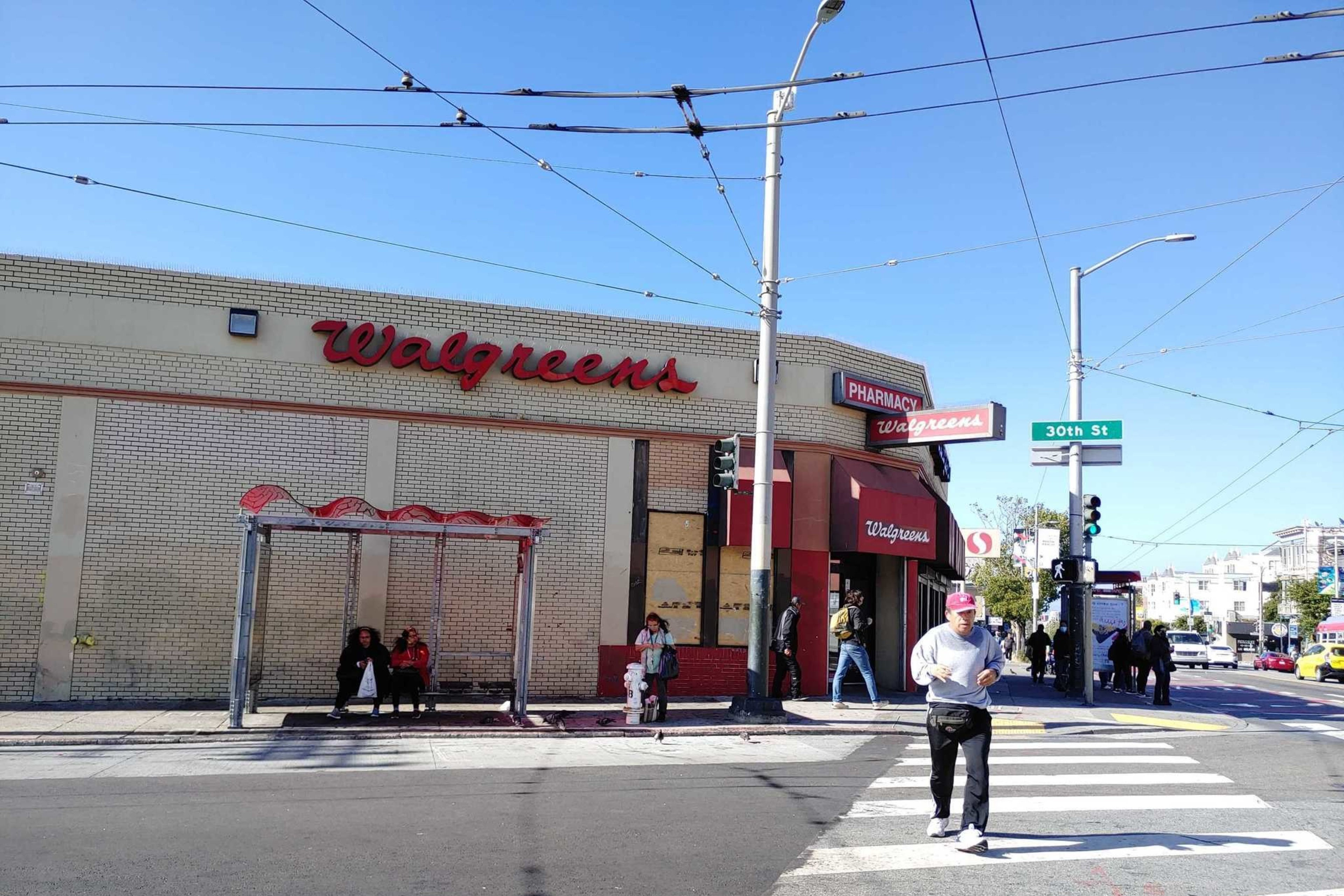 The image shows a Walgreens store on a sunny day, with people walking and sitting at a bus stop. The location is at the corner of a street with a 30th St sign.