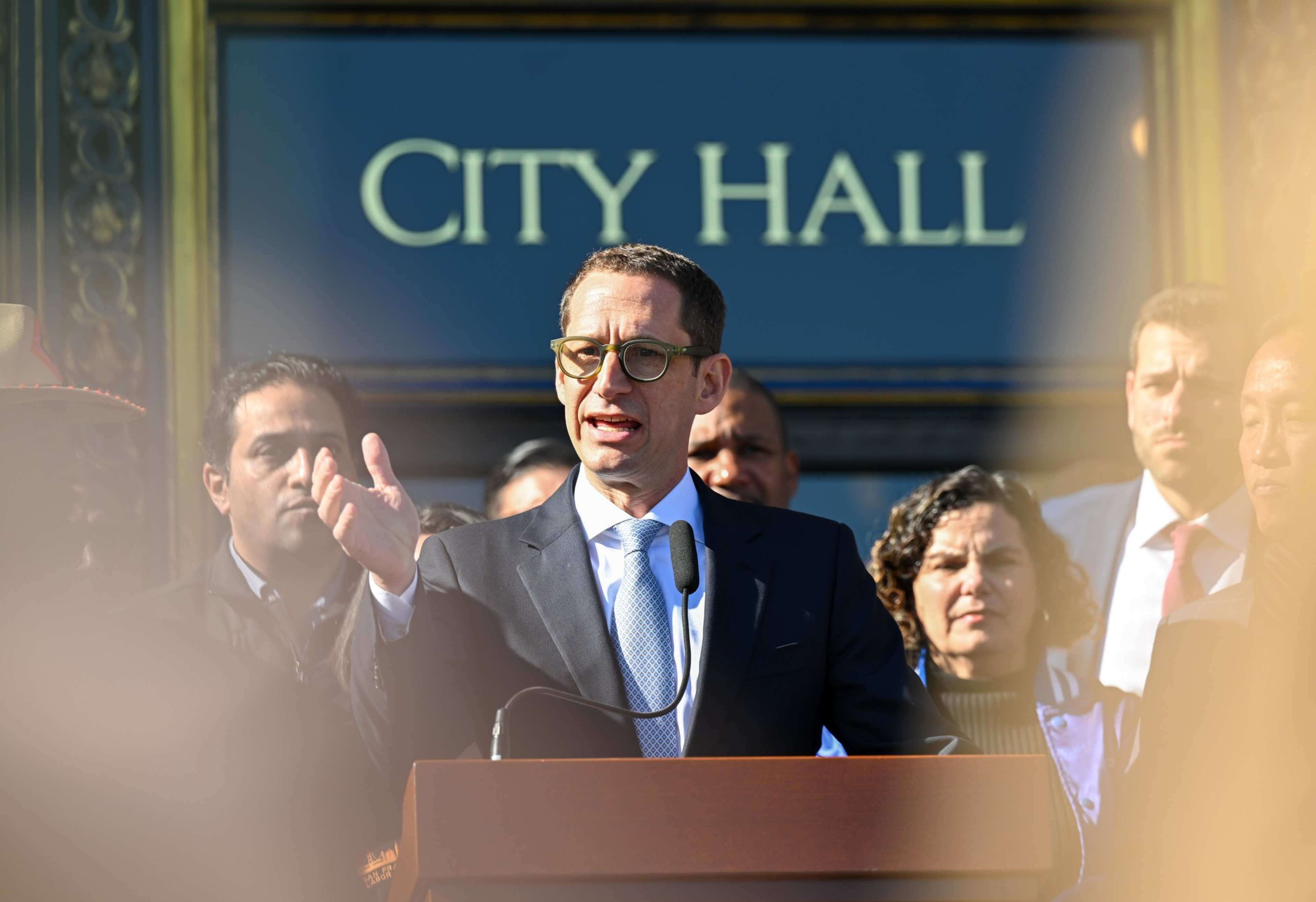 A man in a suit and glasses speaks at a City Hall podium, surrounded by attentive people in formal attire under sunlight.