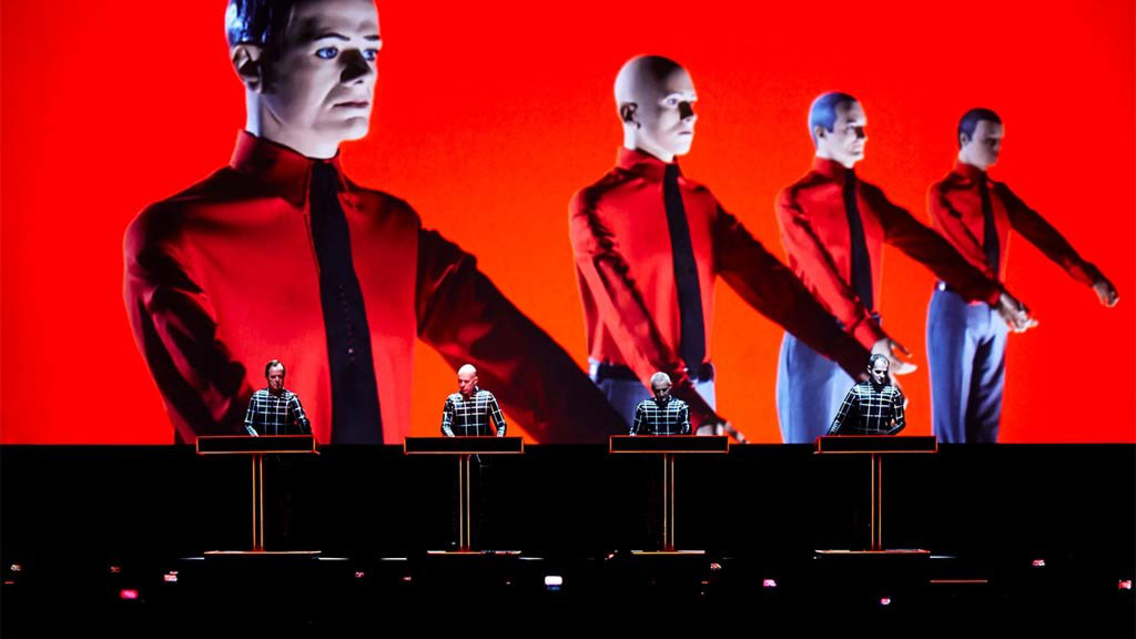 Four men in black grid-patterned shirts stand behind illuminated podiums, with large projections of mannequins in red shirts and black ties behind them.