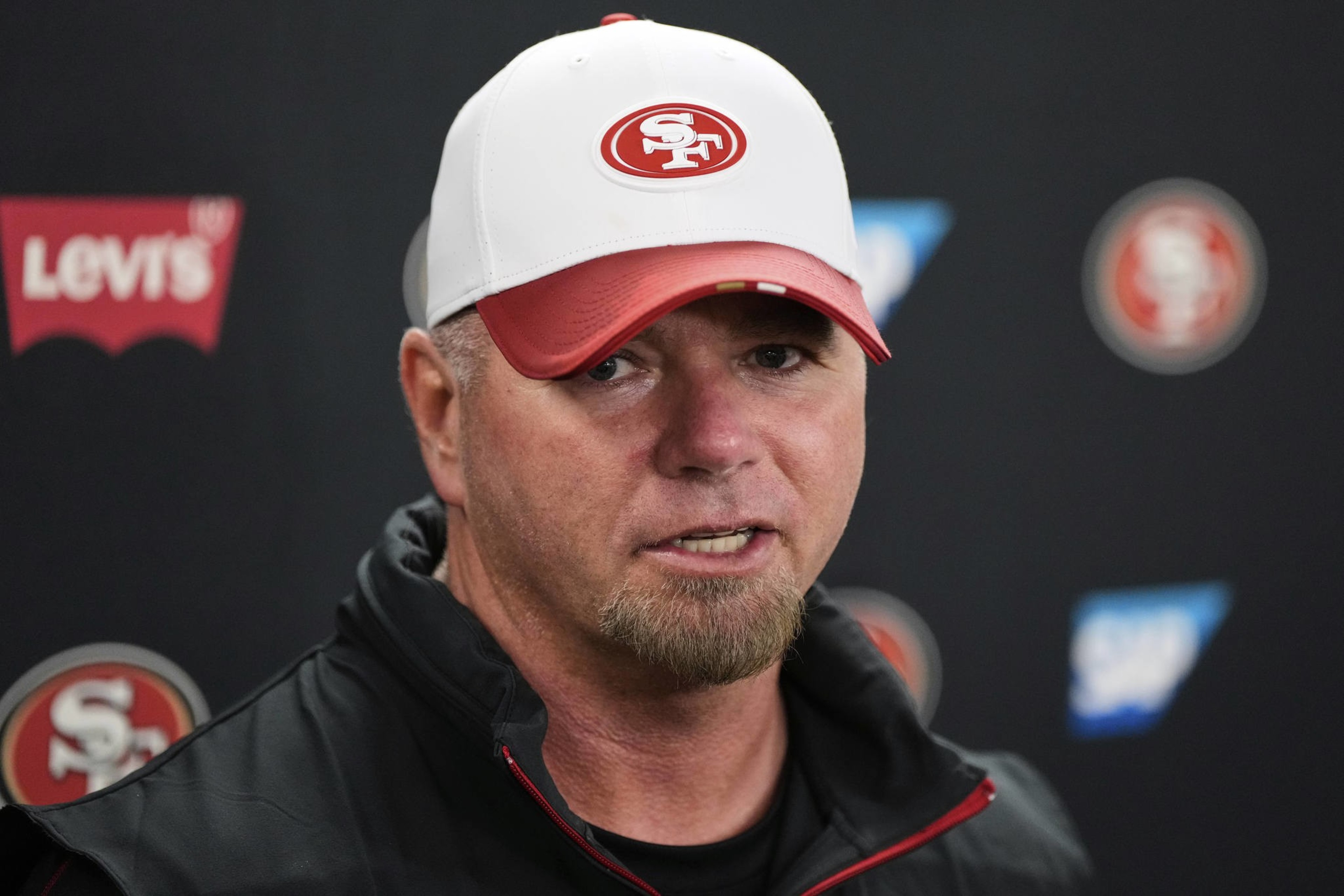 A man in a white and red cap with a &quot;49ers&quot; logo is speaking at a press event. The background features &quot;Levi's&quot; and &quot;SAP&quot; logos.