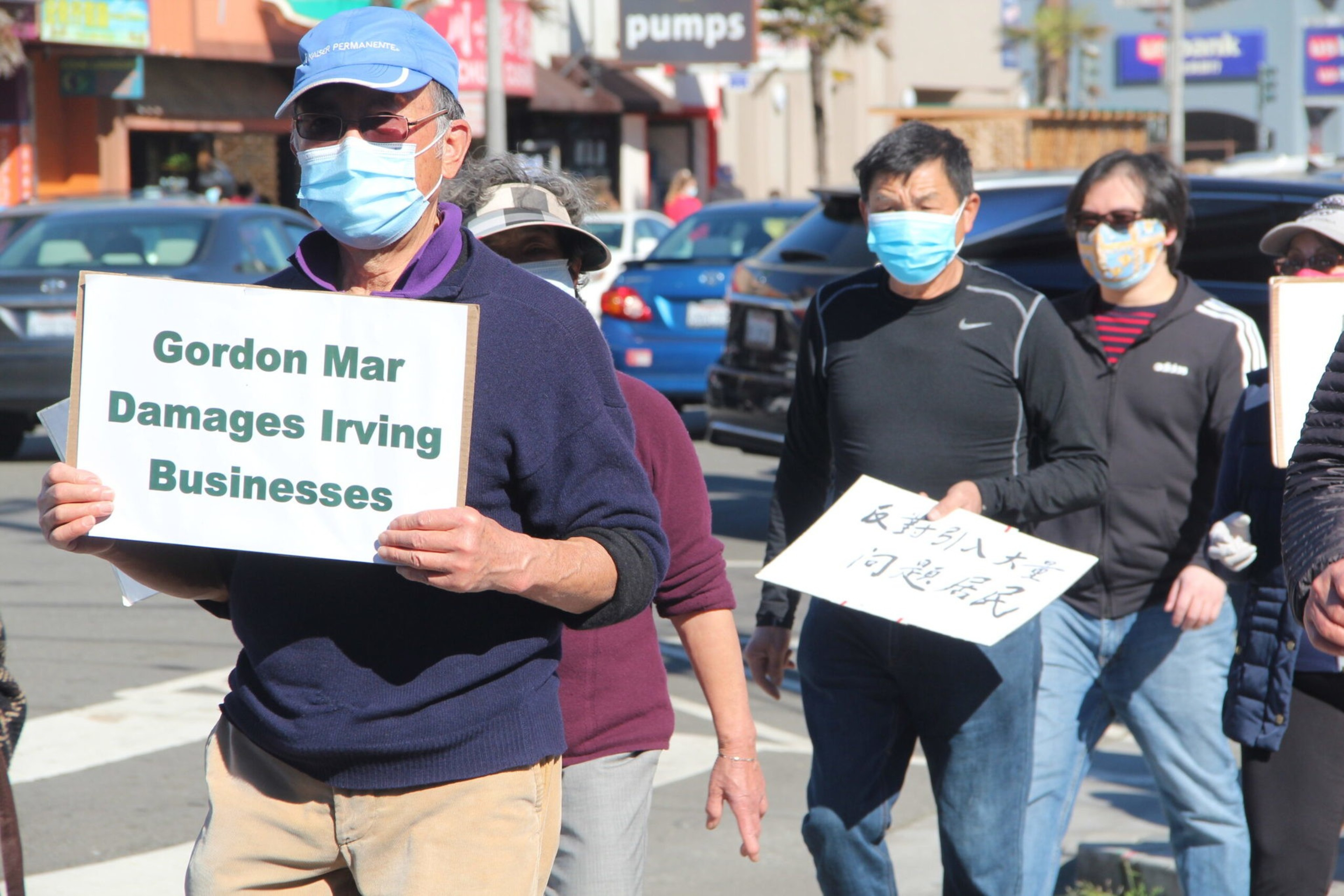 People wearing face masks hold protest signs, with one sign reading "Gordon Mar Damages Irving Businesses," as they walk along a street.