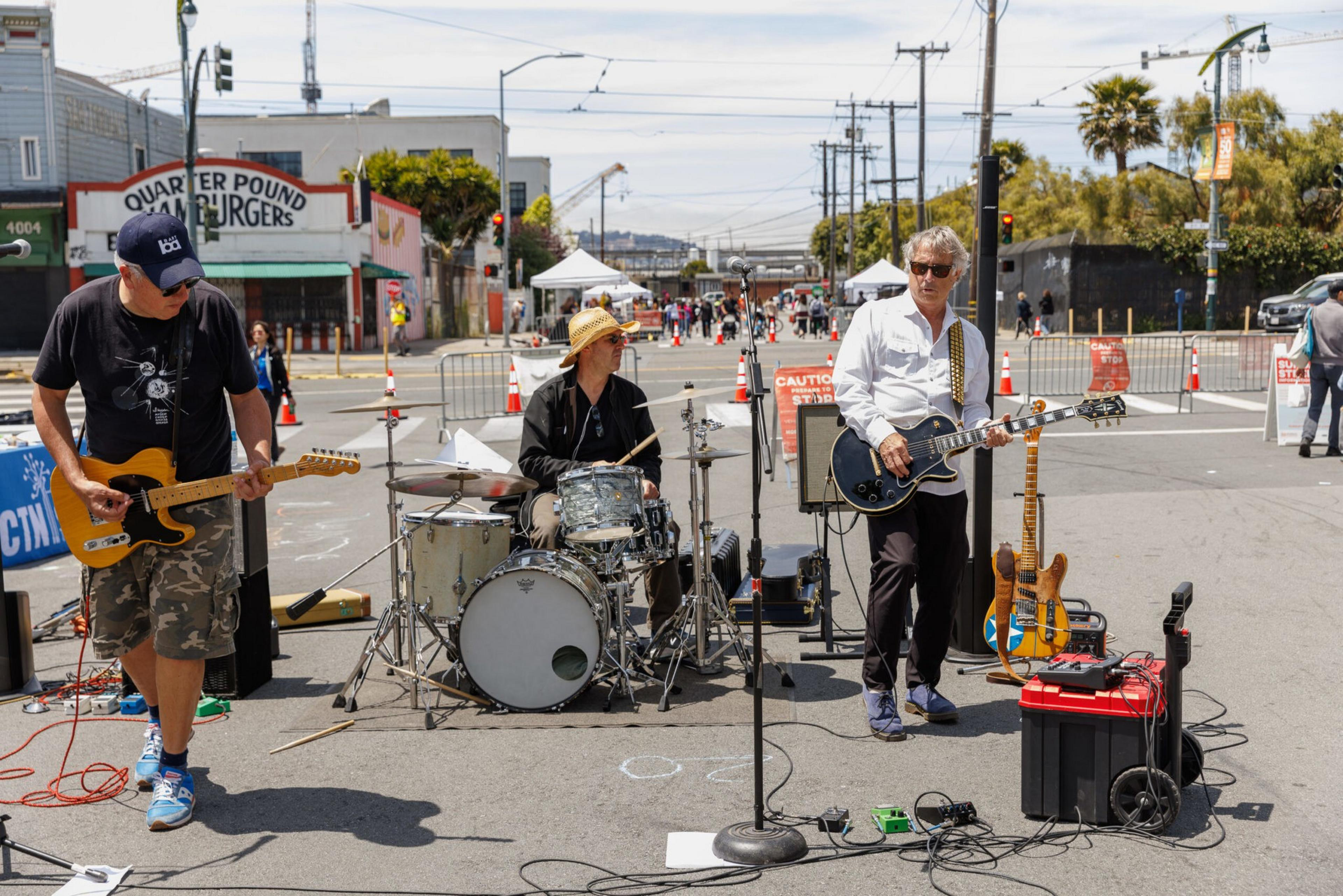 A band with three members plays on a sunny street: two guitarists and a drummer.