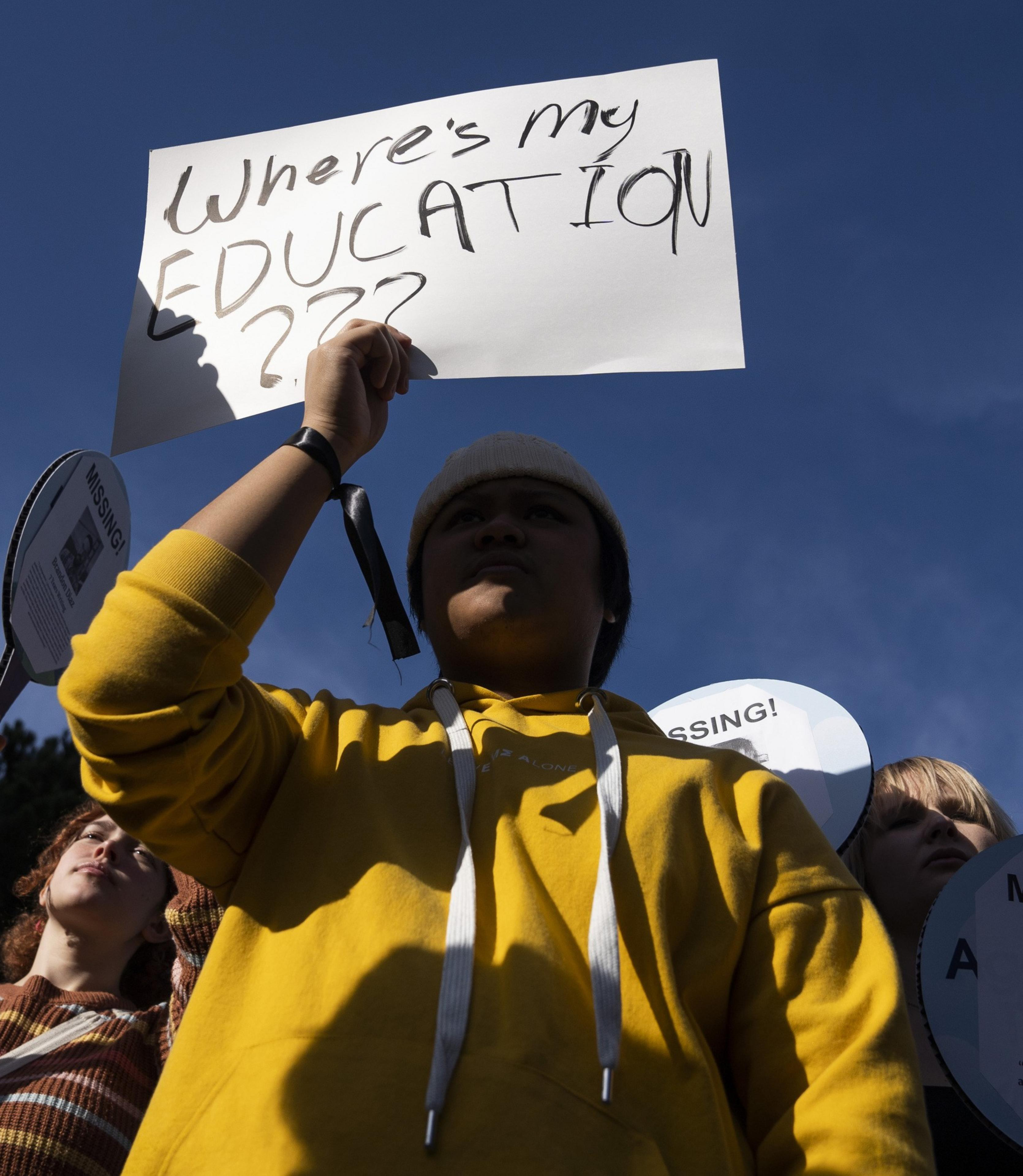 A person in a yellow hoodie holds a sign reading &quot;Where's my EDUCATION???&quot; during a protest, with others in the background under a clear blue sky.